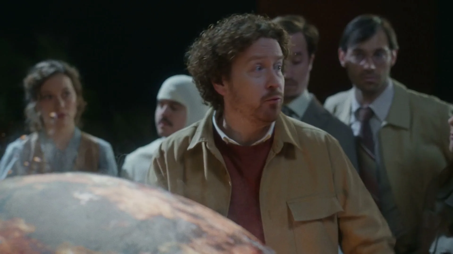 A man with curly hair and a tan jacket looks surprised and points, surrounded by other people in a dimly lit, indeterminate setting.