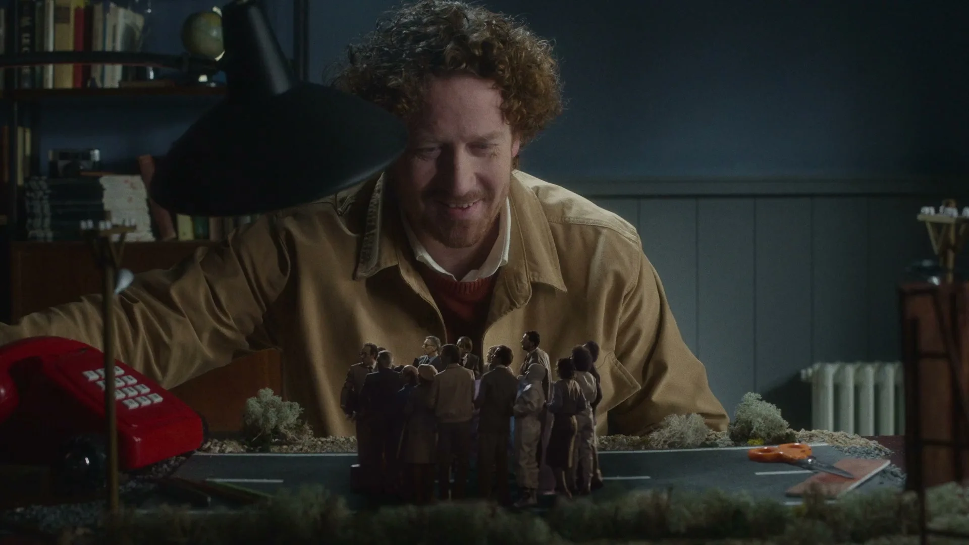 A man with curly red hair smiles down at a miniature diorama of people on a road, with a red phone in the foreground.