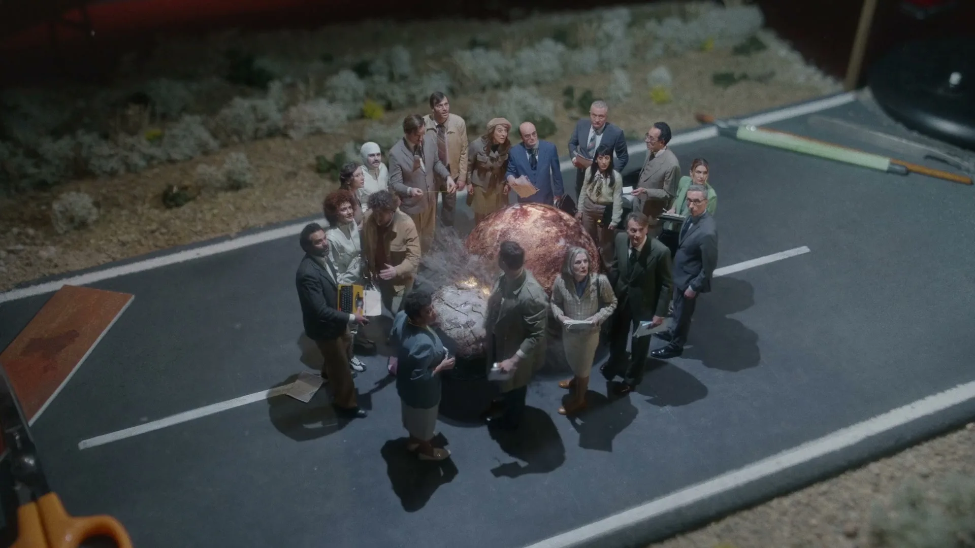 A group of diverse people in retro clothing gather around a miniature, smoking, cracked planet in a diorama of a desert road.