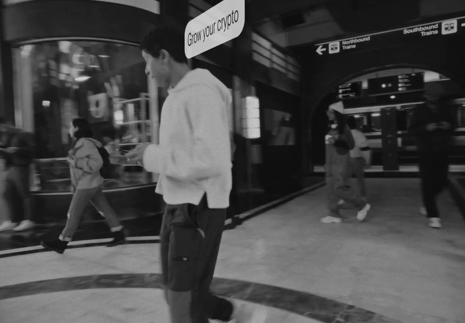 A young man in a hoodie walks through a busy, black and white subway station, looking at his phone, as a thought bubble appears above his head.
