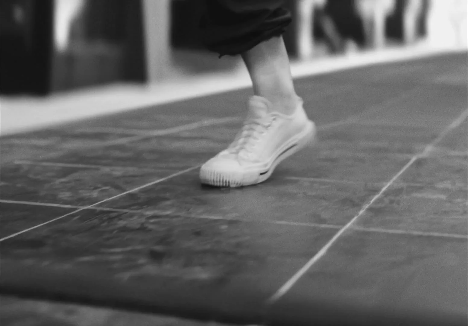 A person wearing white sneakers and dark pants walks forward, stepping onto a raised platform in a black and white shot.