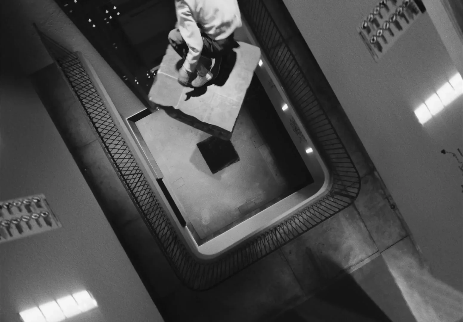 A person in a white hoodie and dark pants jumps onto a floating platform in a dark, multi-story building interior, shot in black and white.