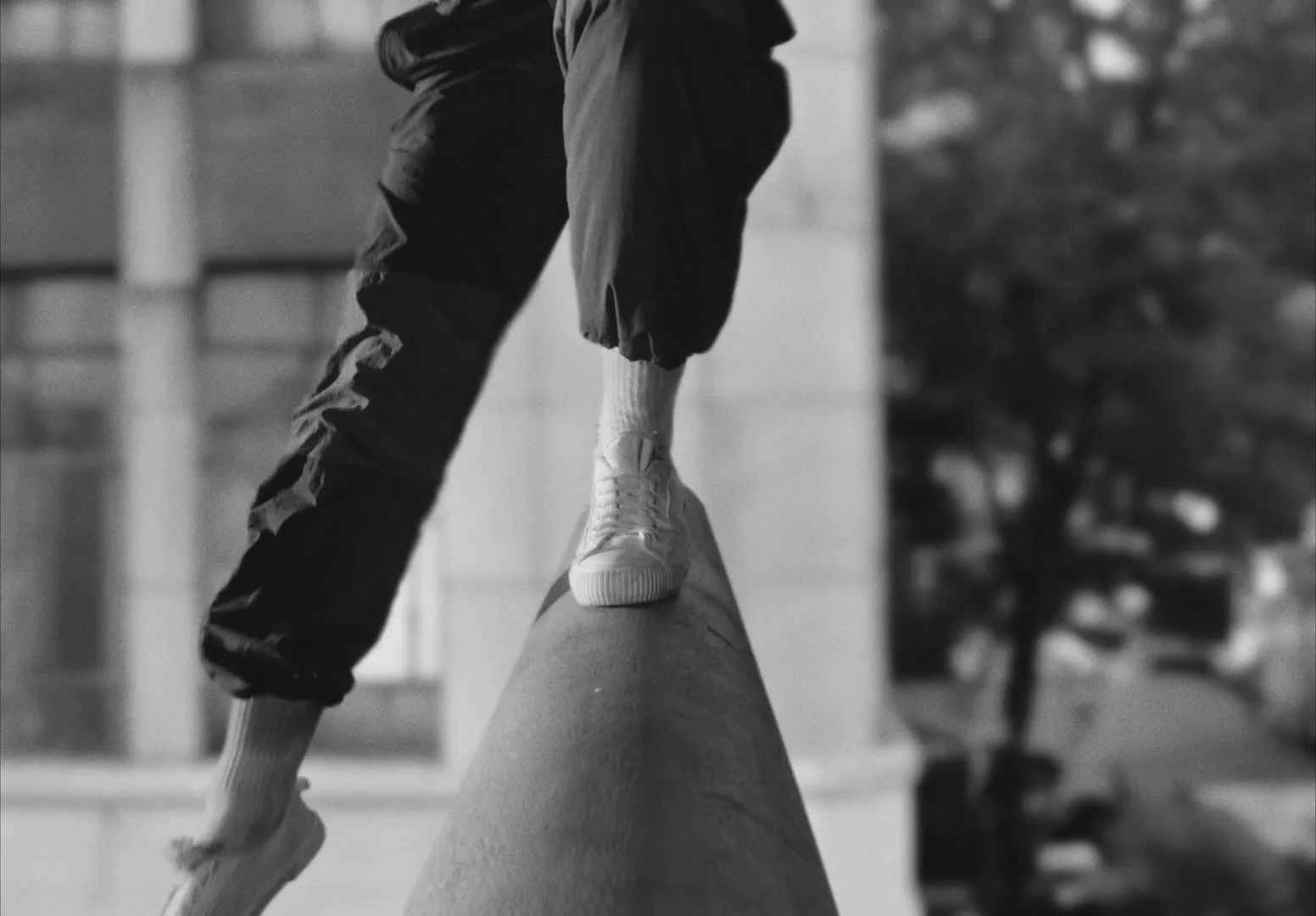 A person in dark pants and white sneakers balances on a narrow concrete ledge in a city setting.