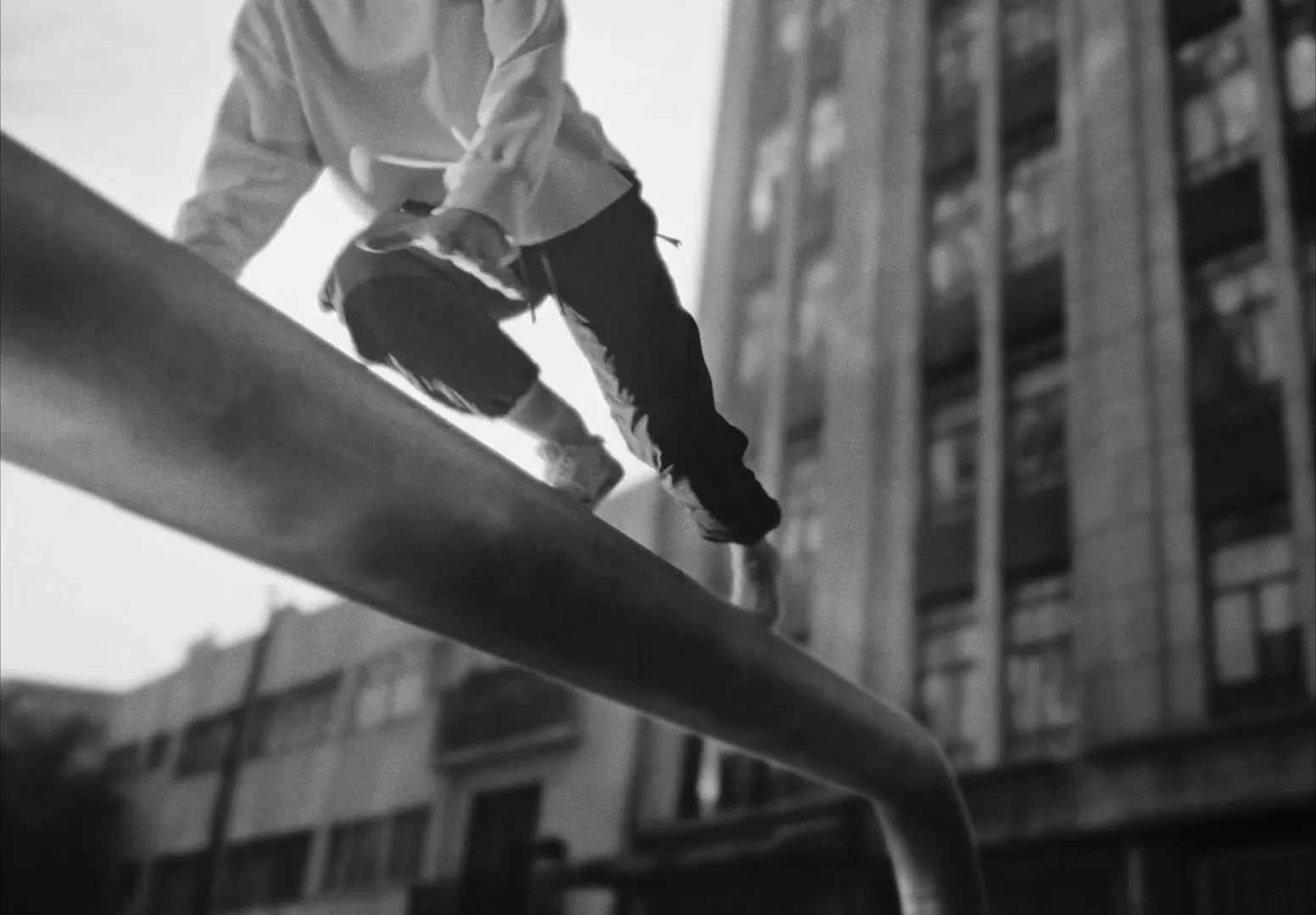 A young man in a hoodie and pants balances on a large metal pipe, performing parkour in an urban setting with buildings in the background.