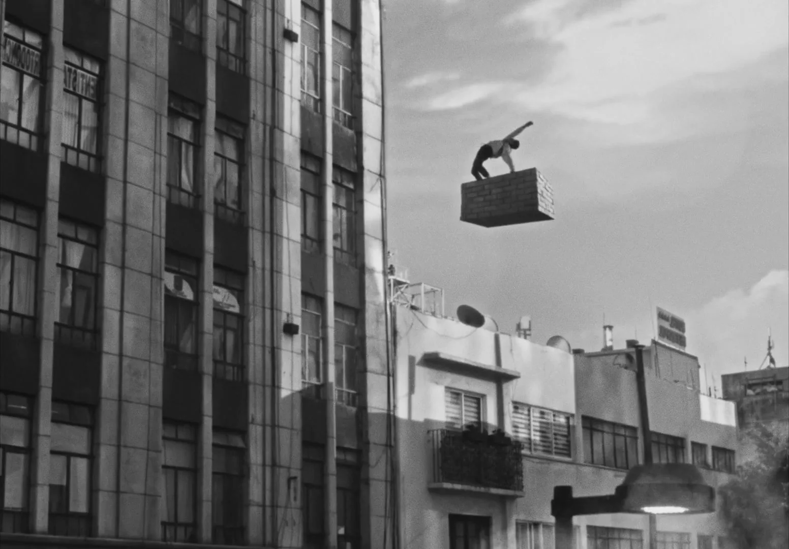 A man in a tank top balances on a floating brick platform, performing acrobatics against a cloudy sky above city buildings.