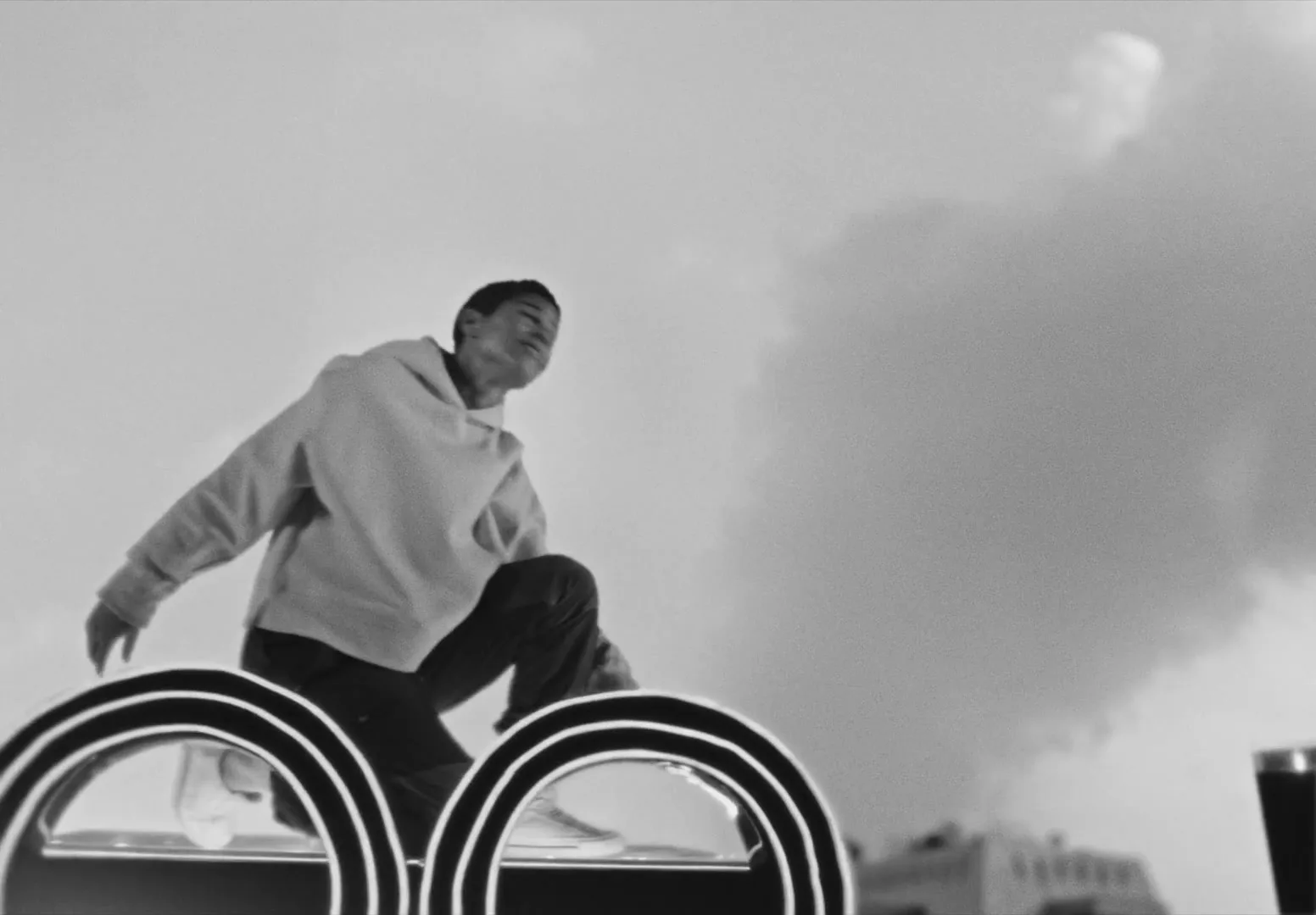 A young man in a hoodie crouches on a rooftop structure, then jumps off, silhouetted against a cloudy sky in a city.