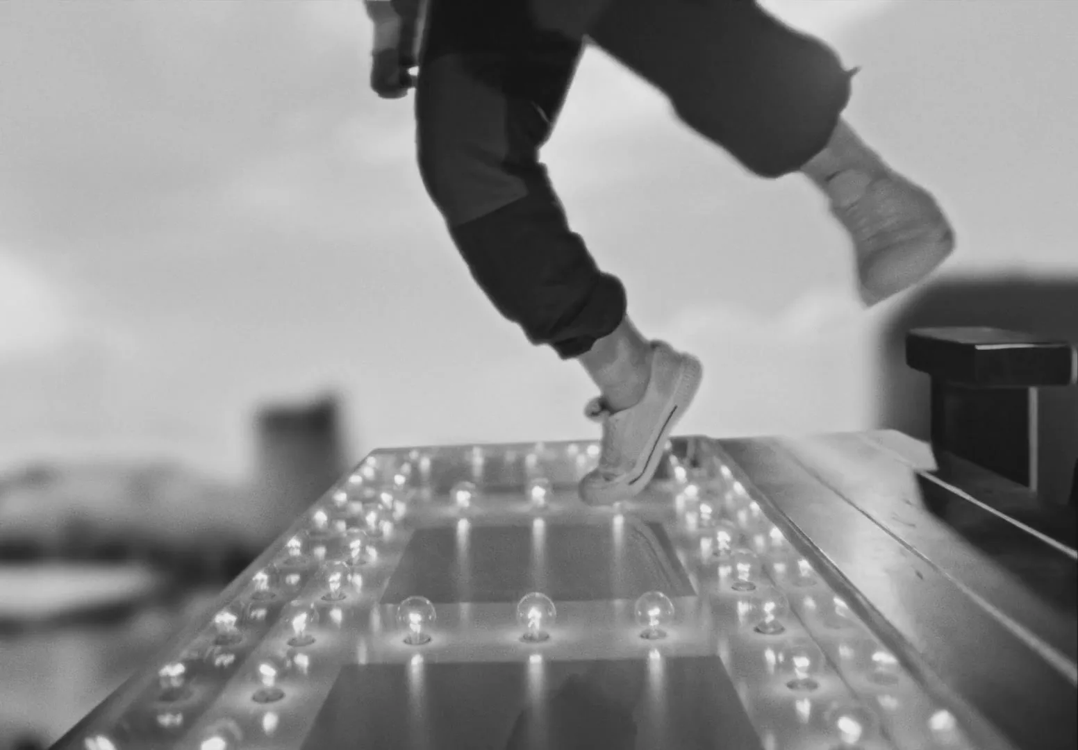 A person in dark pants and light shoes walks on a brightly lit platform, with a blurred background of a city skyline under a bright sky.