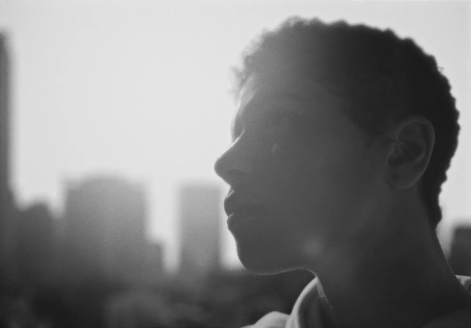 A young person with short curly hair is seen in profile, looking up and to the right, with a bright, hazy city skyline in the background.