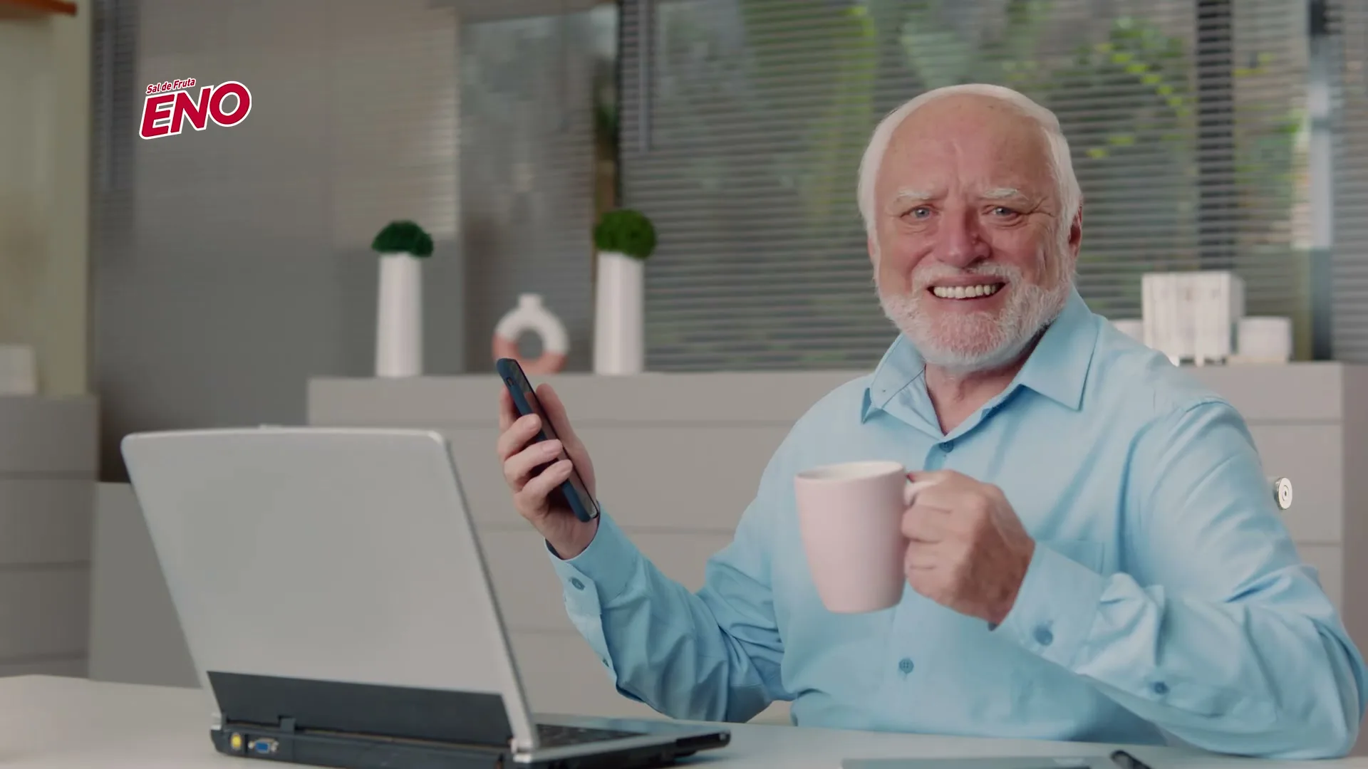 An older man with white hair and a beard, wearing a blue shirt, smiles awkwardly at the camera while holding a phone and a mug at a desk with a laptop.