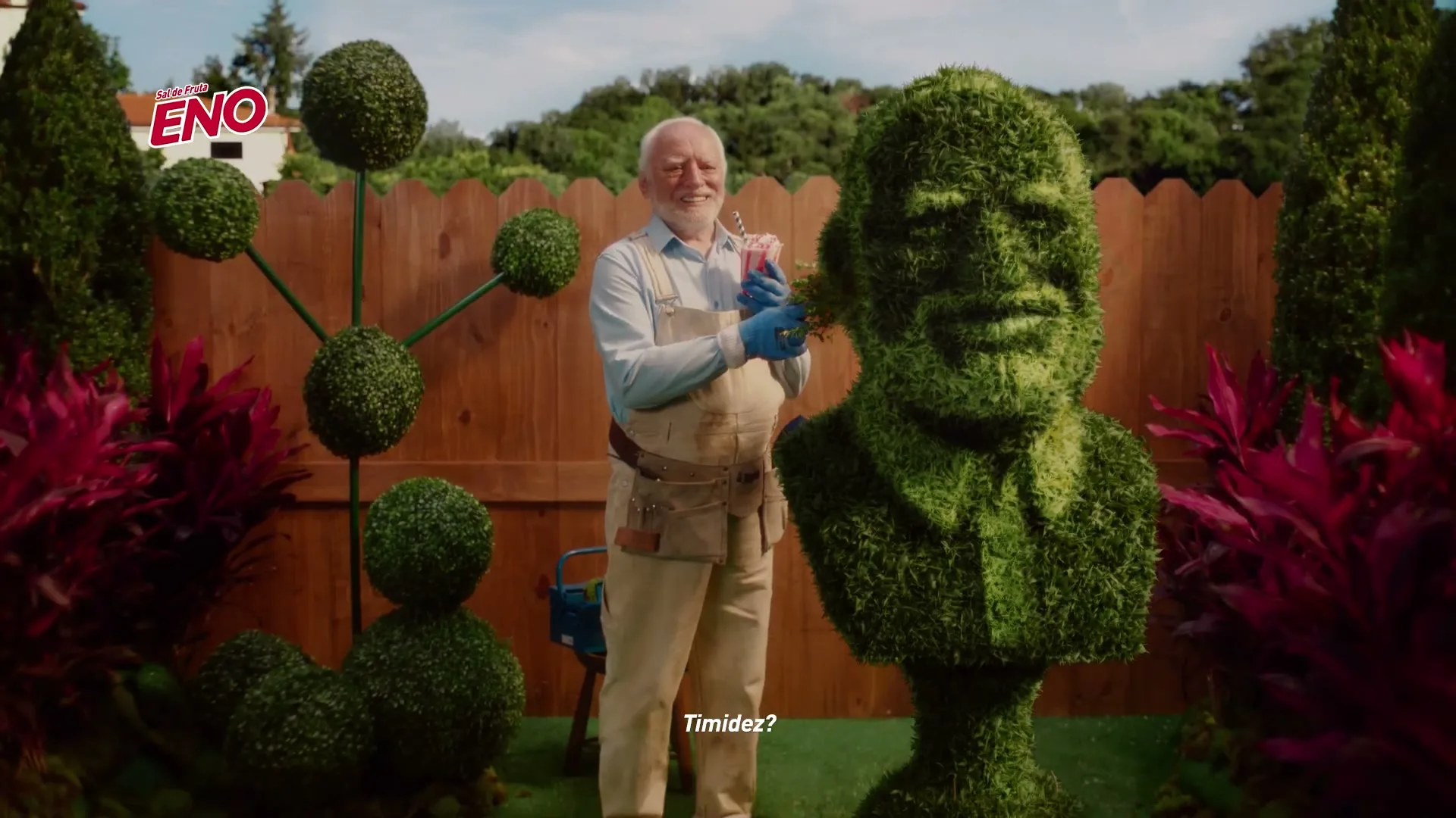 An older man in gardening overalls smiles while trimming a topiary bust in a sunny garden, holding a pink drink.