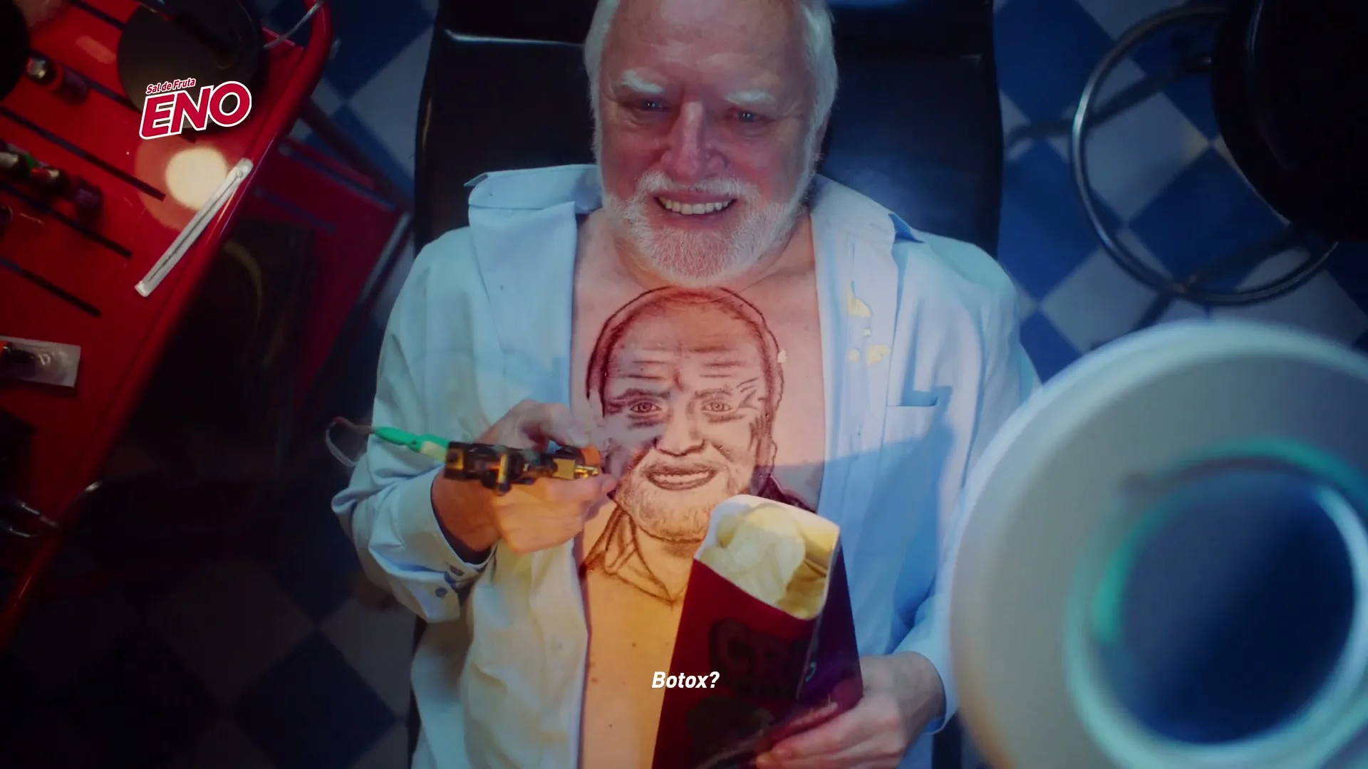 A man with white hair and a blue shirt smiles while getting a tattoo of his own face on his chest in a tattoo parlor.