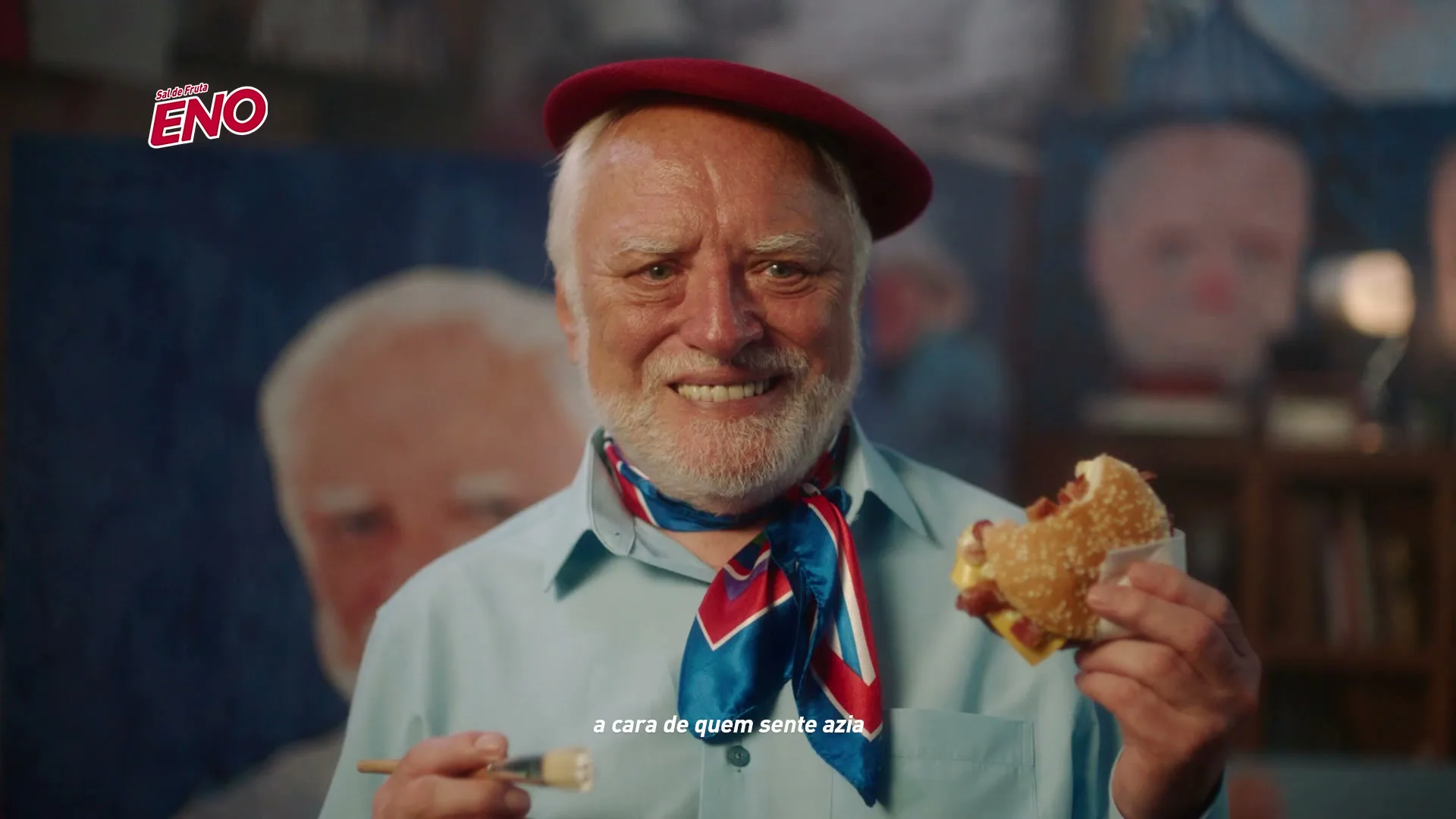 An older man with a red beret and a blue scarf smiles awkwardly while holding a half-eaten burger and a paintbrush in an art studio.