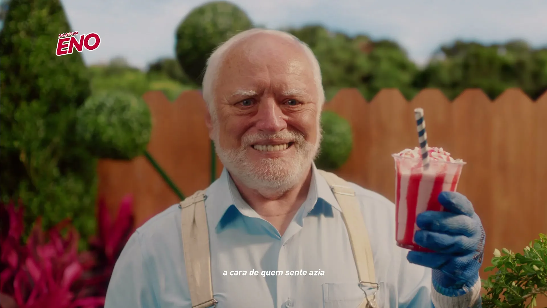An older man with white hair and beard, wearing a light blue shirt and suspenders, smiles awkwardly while holding a red and white drink in a garden.