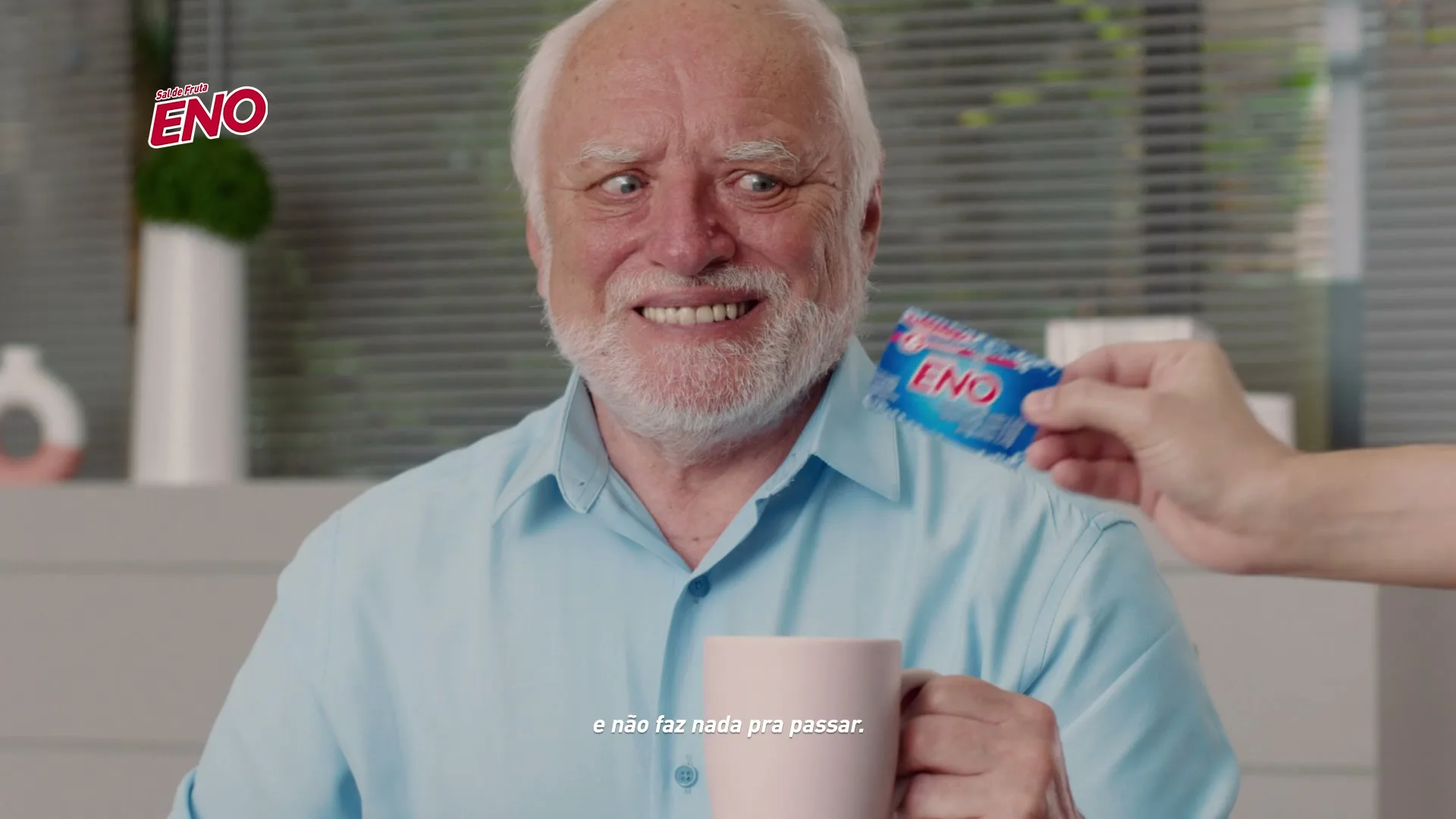 An older man with a white beard and blue shirt holds a pink mug, looking uncomfortable as a hand offers him a small blue packet of Eno.