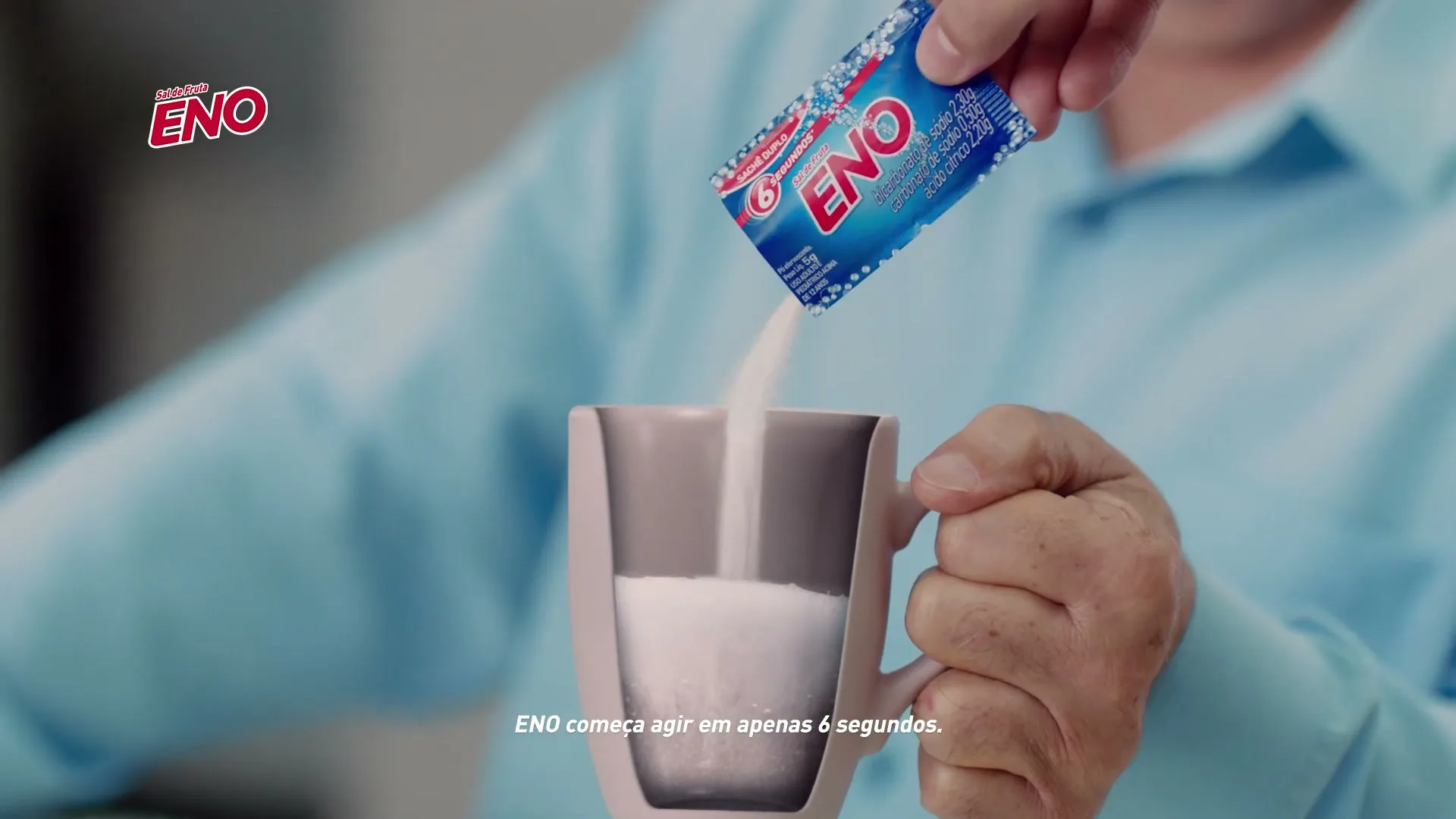 A person in a light blue shirt holds a pink mug while pouring white powder from a blue Eno sachet into it, in an interior setting.