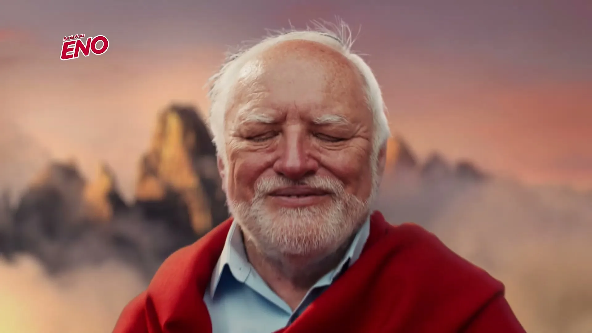 A man with white hair and beard, wearing a blue shirt and red shawl, smiles with closed eyes against a mountain backdrop at sunset.