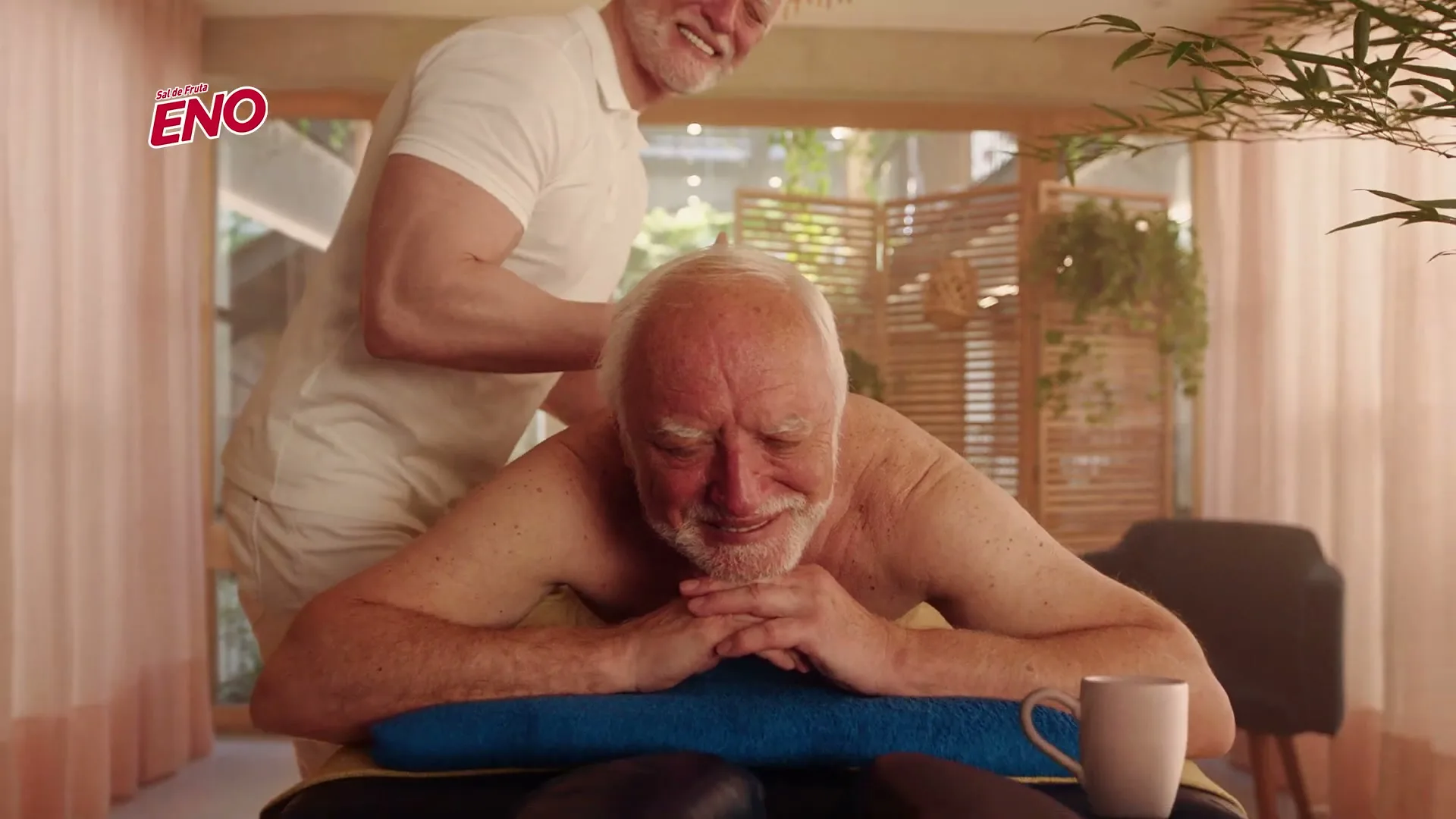 An older man with a white beard lies face down on a massage table, receiving a massage from another man in a bright, airy spa setting.