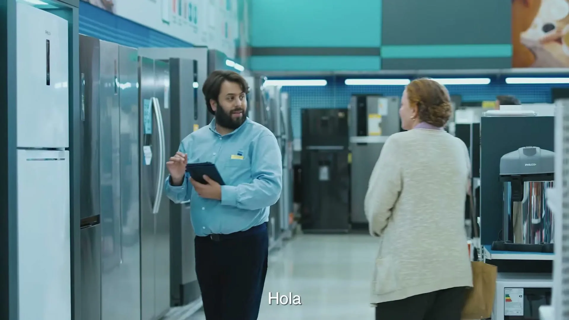 A male salesperson in a blue shirt looks at a tablet while a female customer in a beige cardigan approaches him in an appliance store.