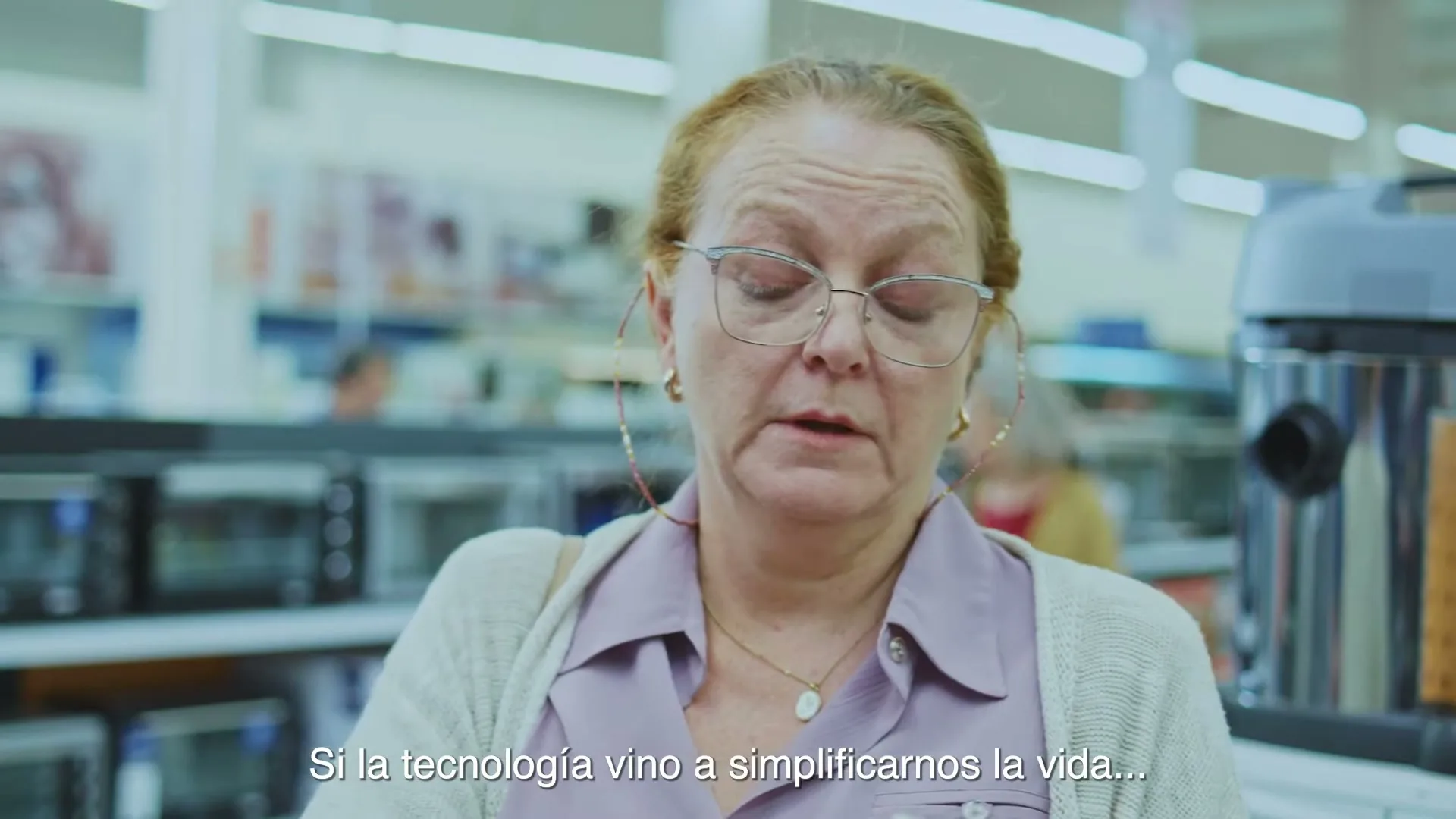 A middle-aged woman with glasses and a light purple shirt looks around a brightly lit electronics store, appearing confused.