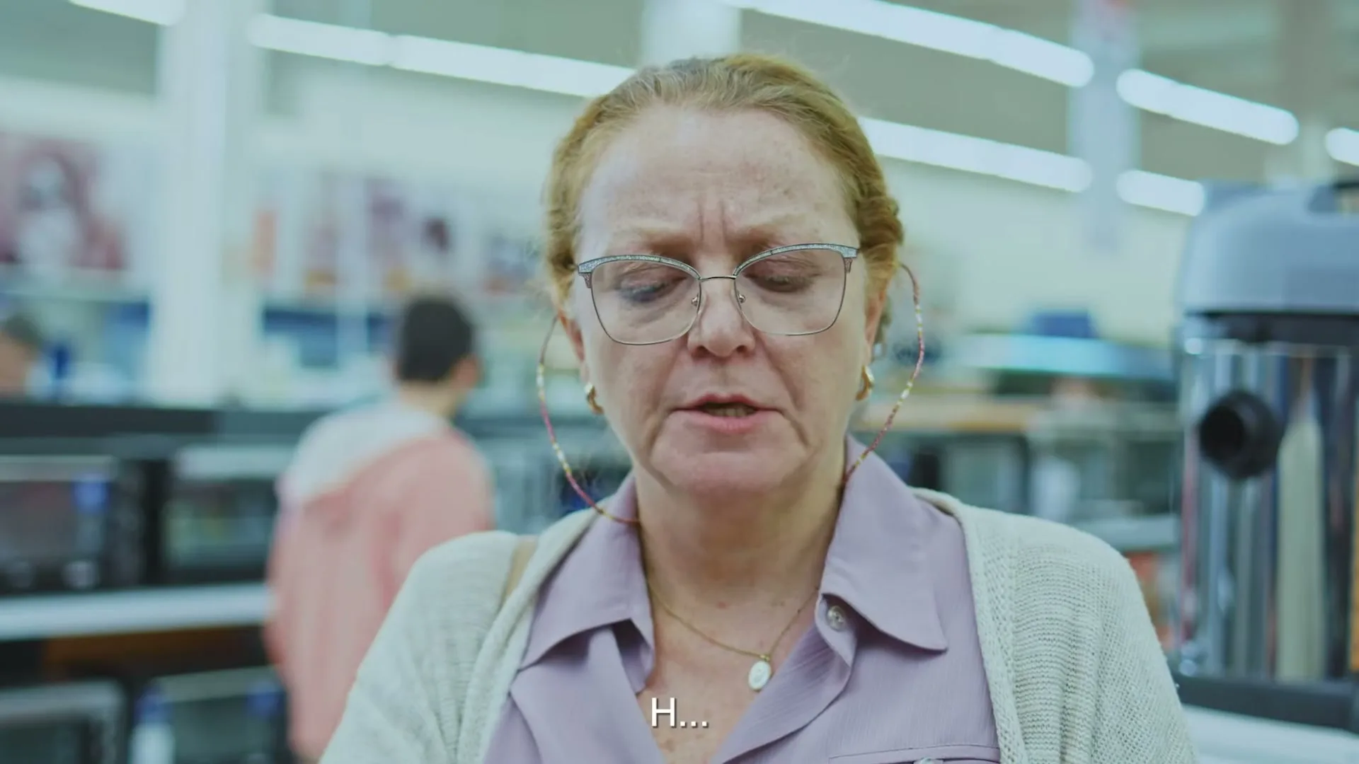 A middle-aged woman with red hair and glasses looks confused while shopping in an electronics store.
