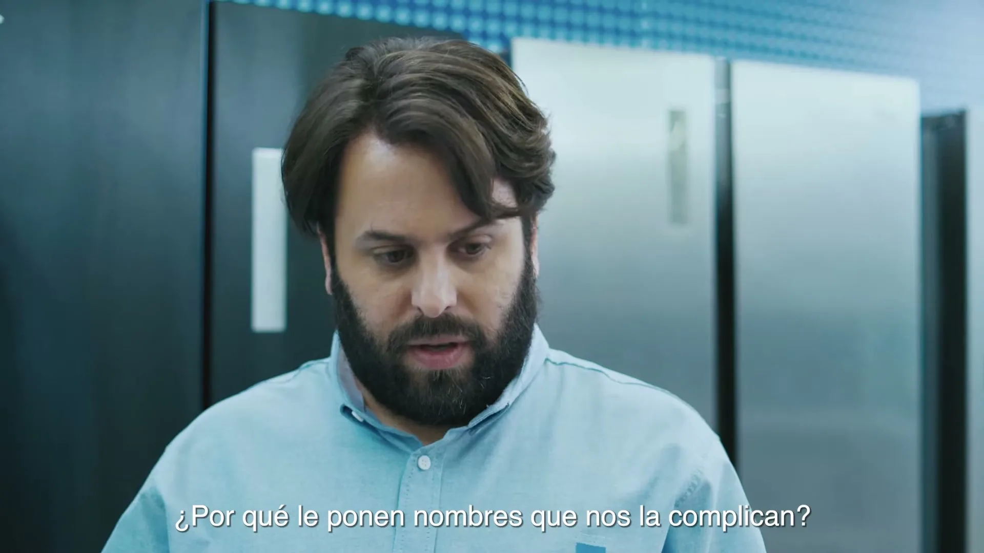 A bearded man in a light blue shirt looks down and then up, speaking in a whisper, in an appliance store with refrigerators in the background.