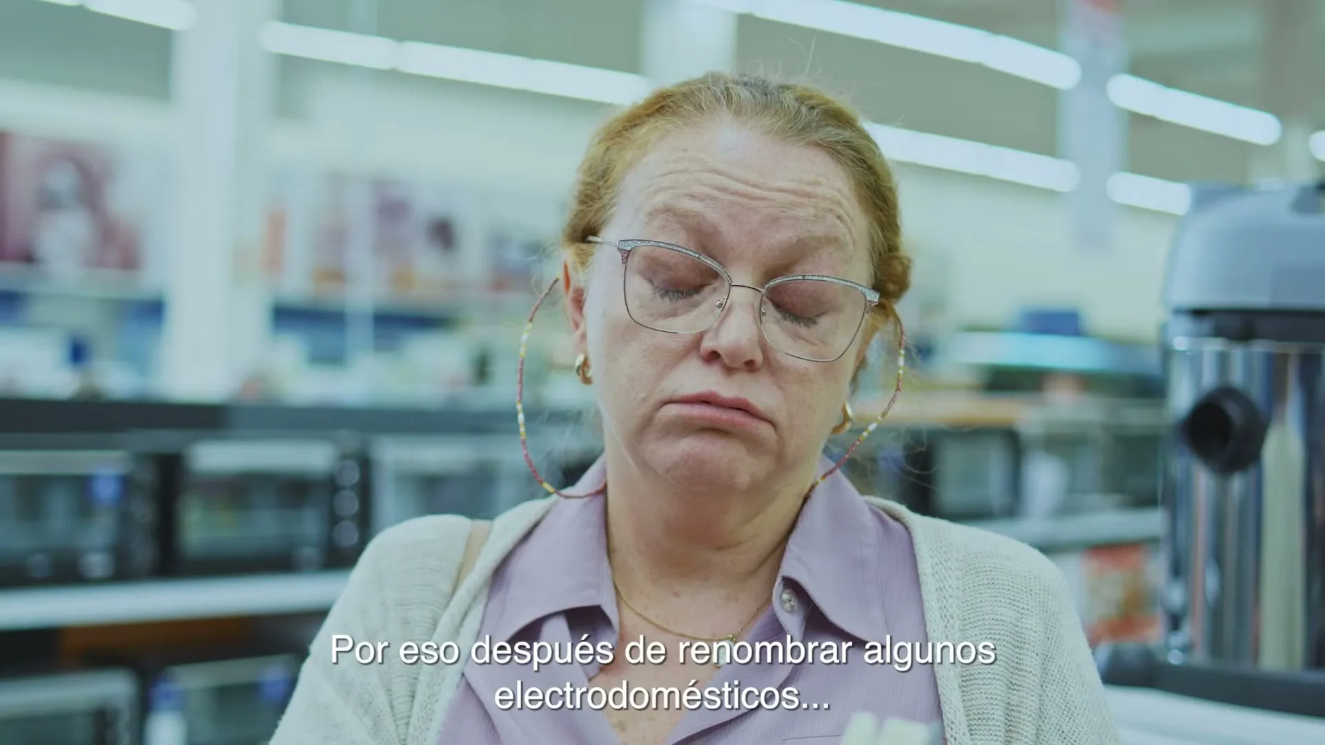 A middle-aged woman with red hair and glasses looks tired and rubs her eyes in an electronics store.