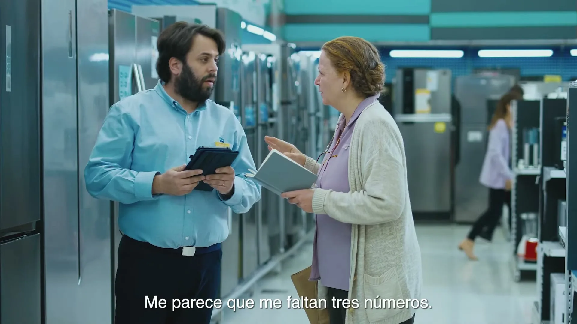 A male store employee in a blue shirt talks to an older female customer in a purple top and cardigan, both holding notebooks, in an appliance store.