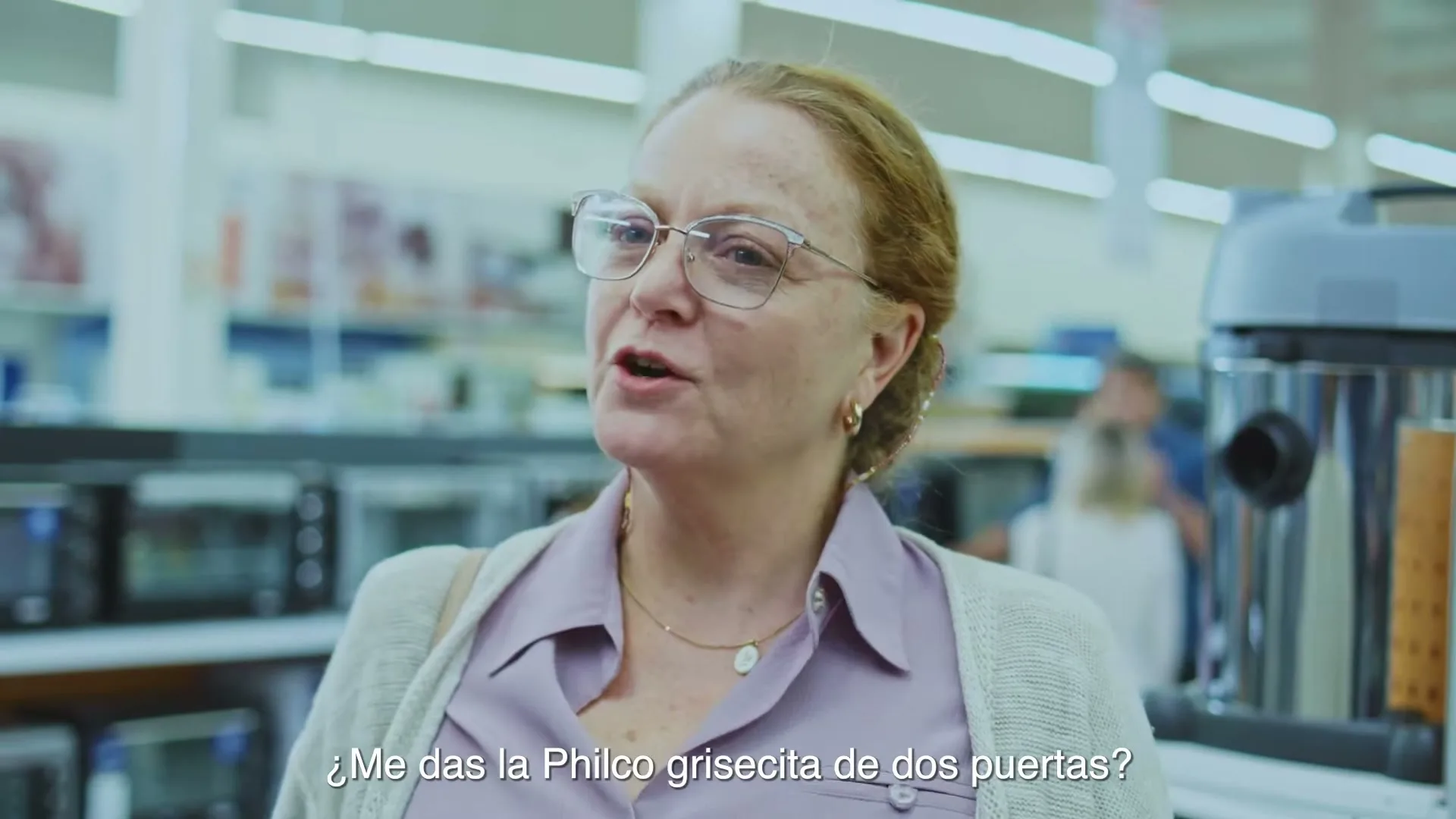 A woman in her 60s, wearing glasses and a light purple shirt, asks for a Philco appliance in an electronics store.