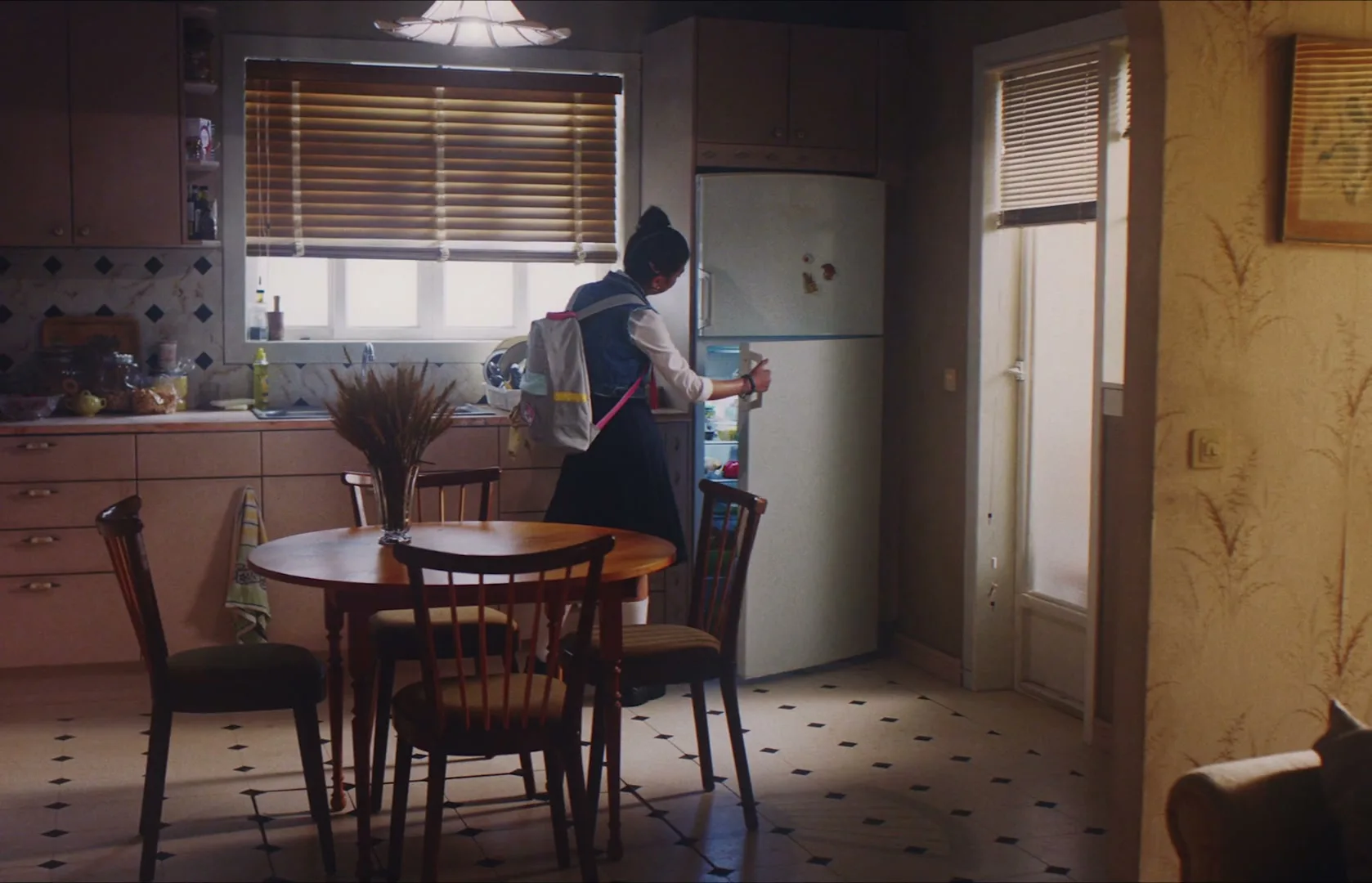 A young female student with a backpack opens a refrigerator in a sunlit kitchen, looking for something to drink.