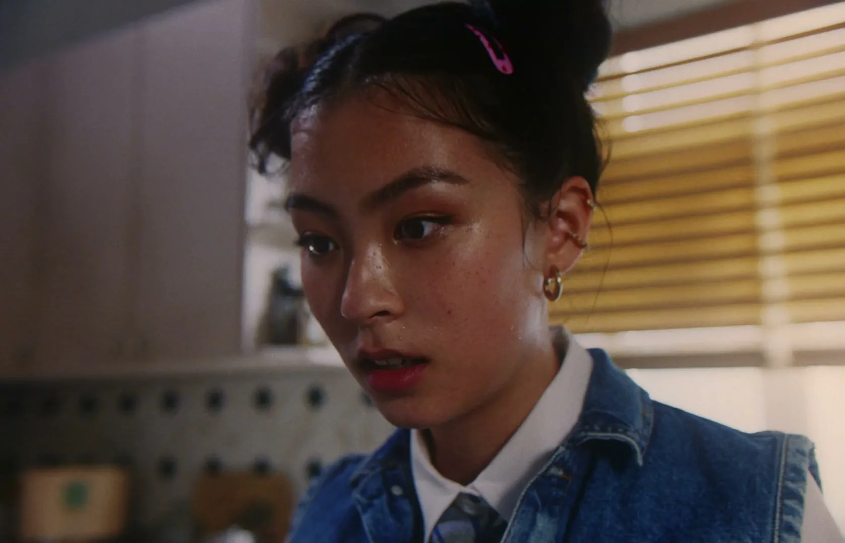 A young woman with space buns and a denim vest looks down with a concerned expression in a kitchen.