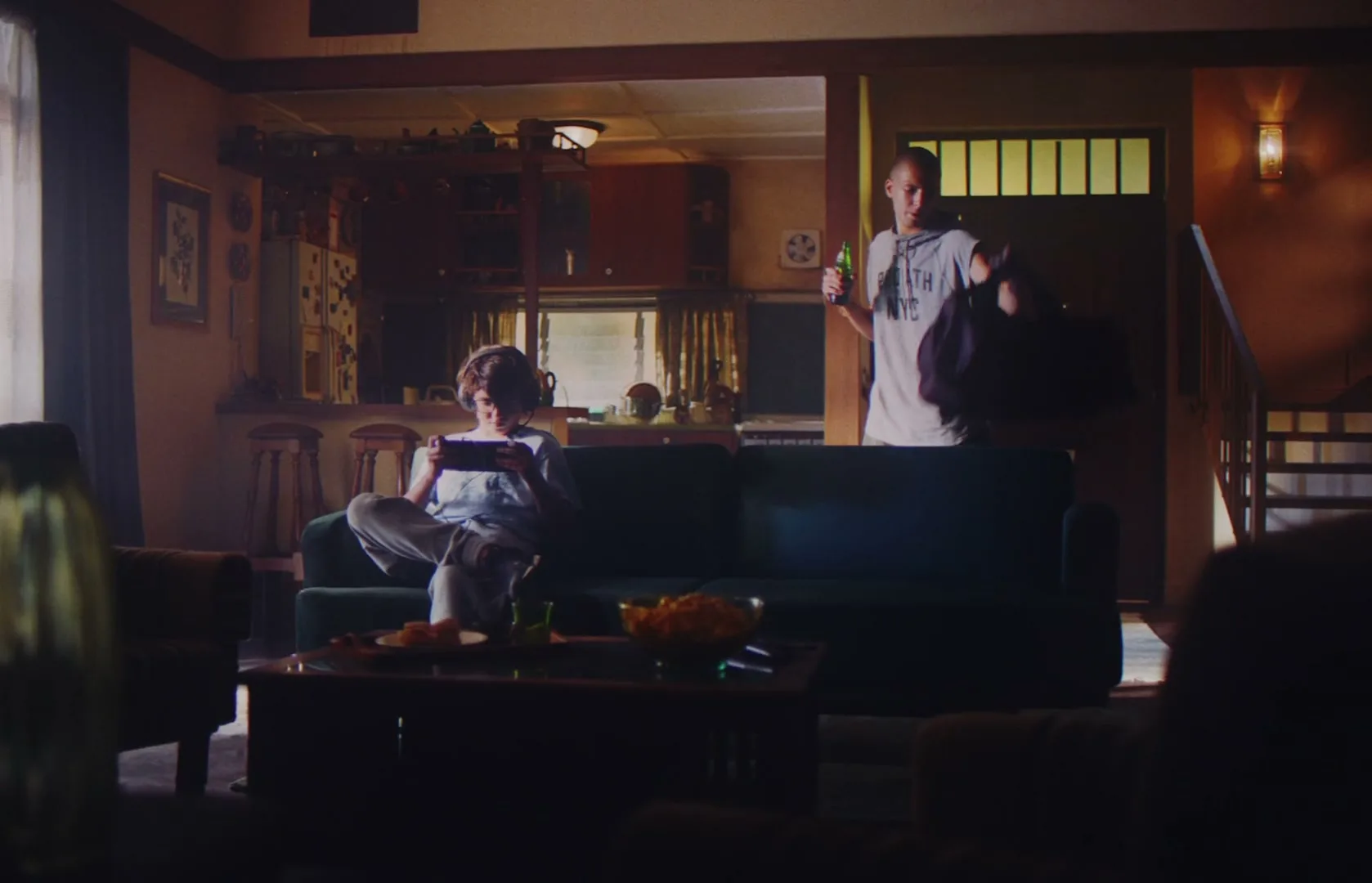A young man in a grey shirt enters a living room with a drink and bag, while another person sits on a couch playing a handheld game.