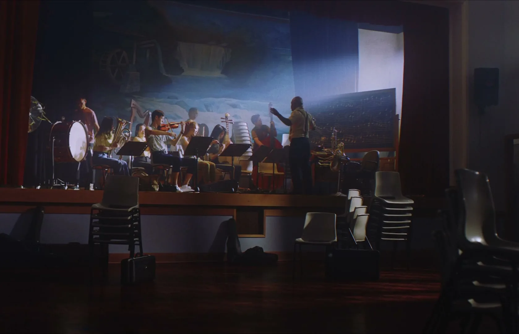 A diverse group of young musicians plays instruments on a stage, led by a conductor, in a dimly lit school auditorium.