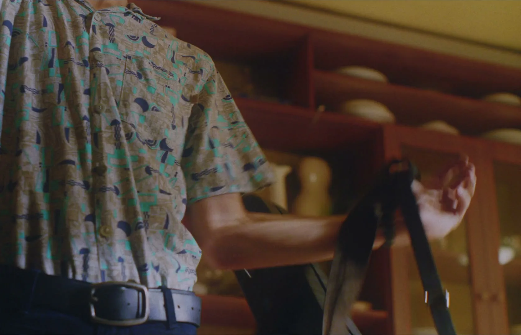 A man in a patterned shirt and dark pants adjusts a bag strap, standing in front of a wooden cabinet with dishes.