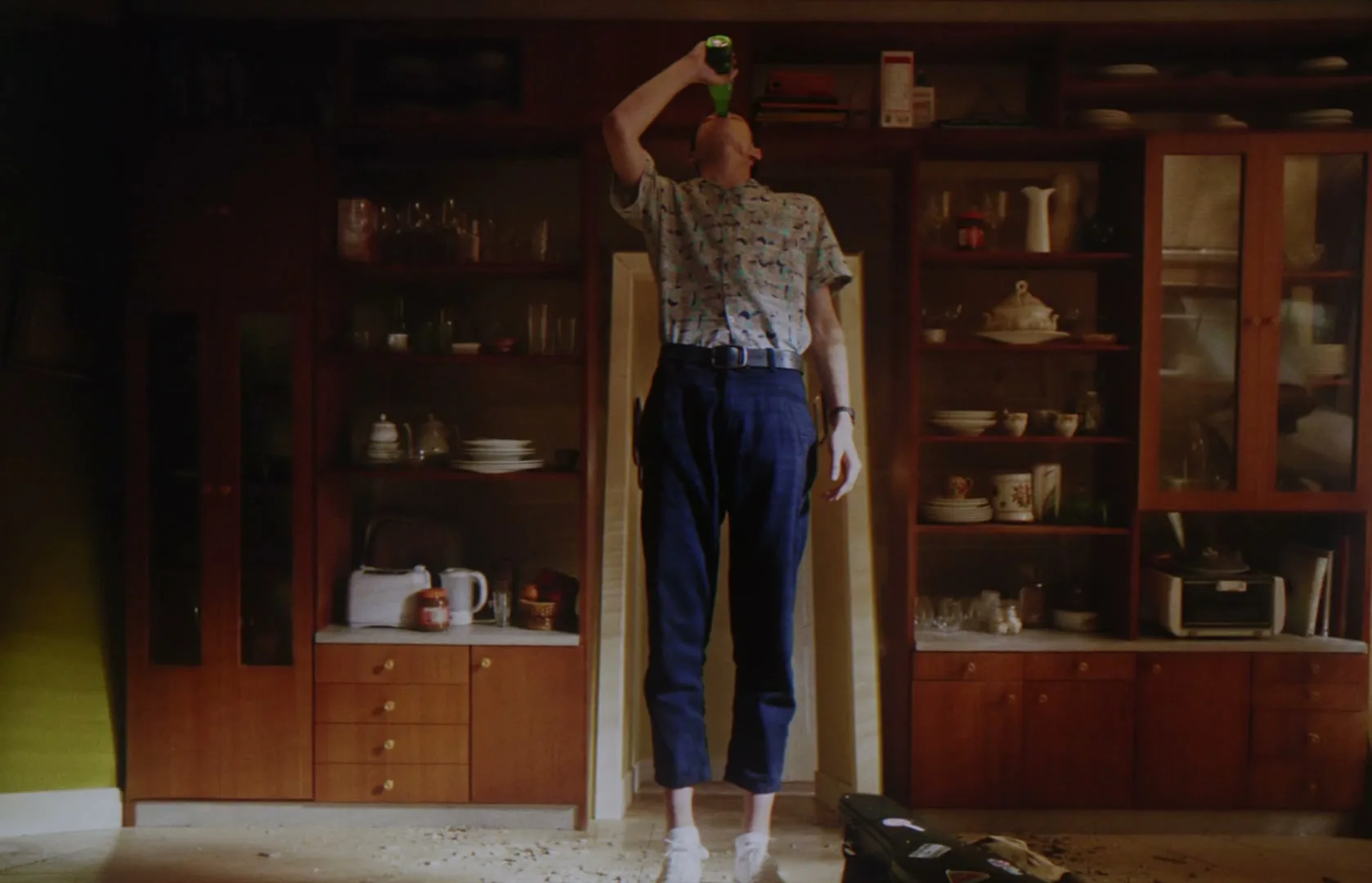 A man in a patterned shirt and blue pants floats in a living room, drinking from a green bottle, as objects fall around him.