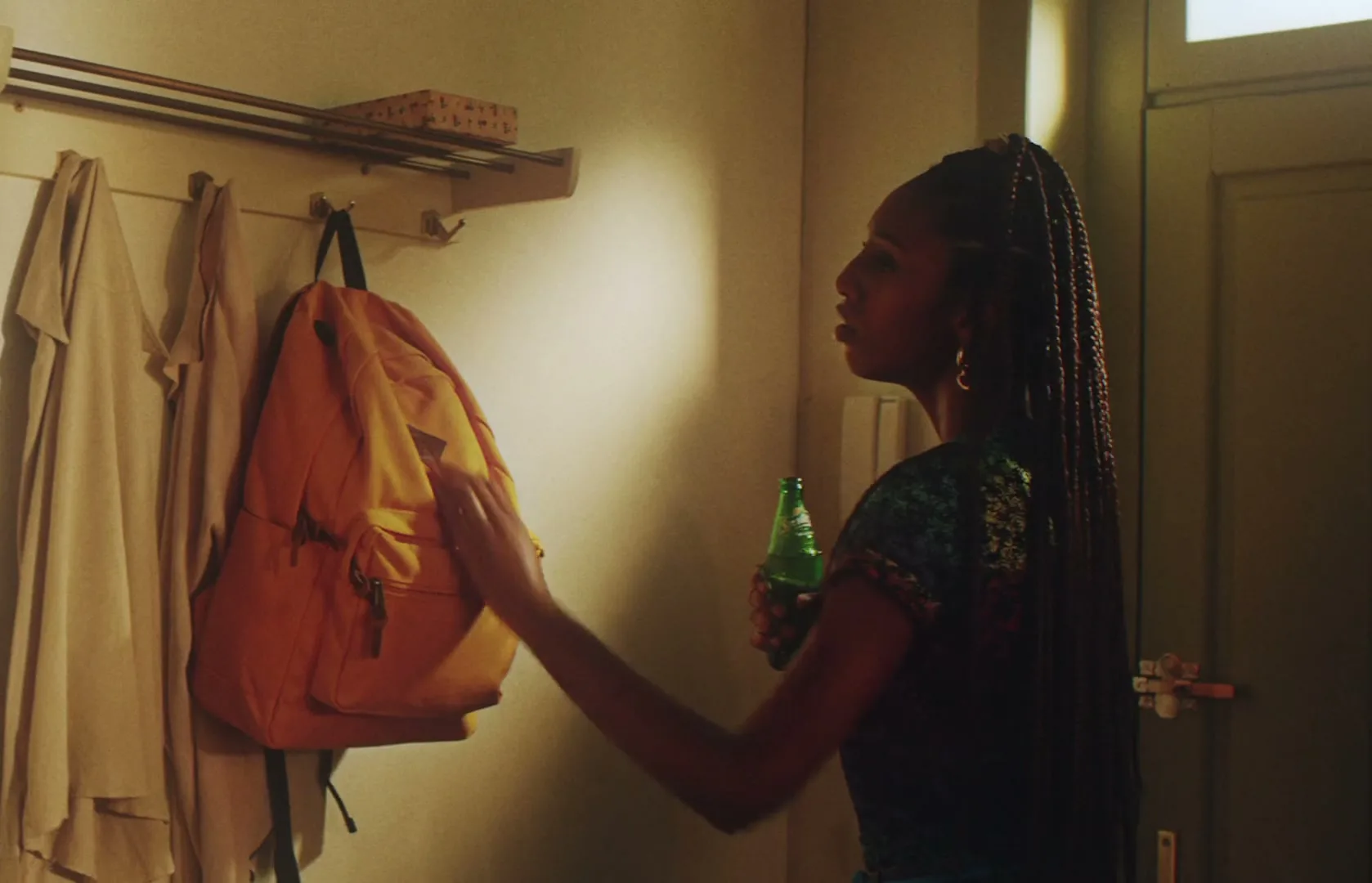 A young woman with long braids hangs her orange backpack on a coat rack, then turns to face the camera, holding a green bottle.