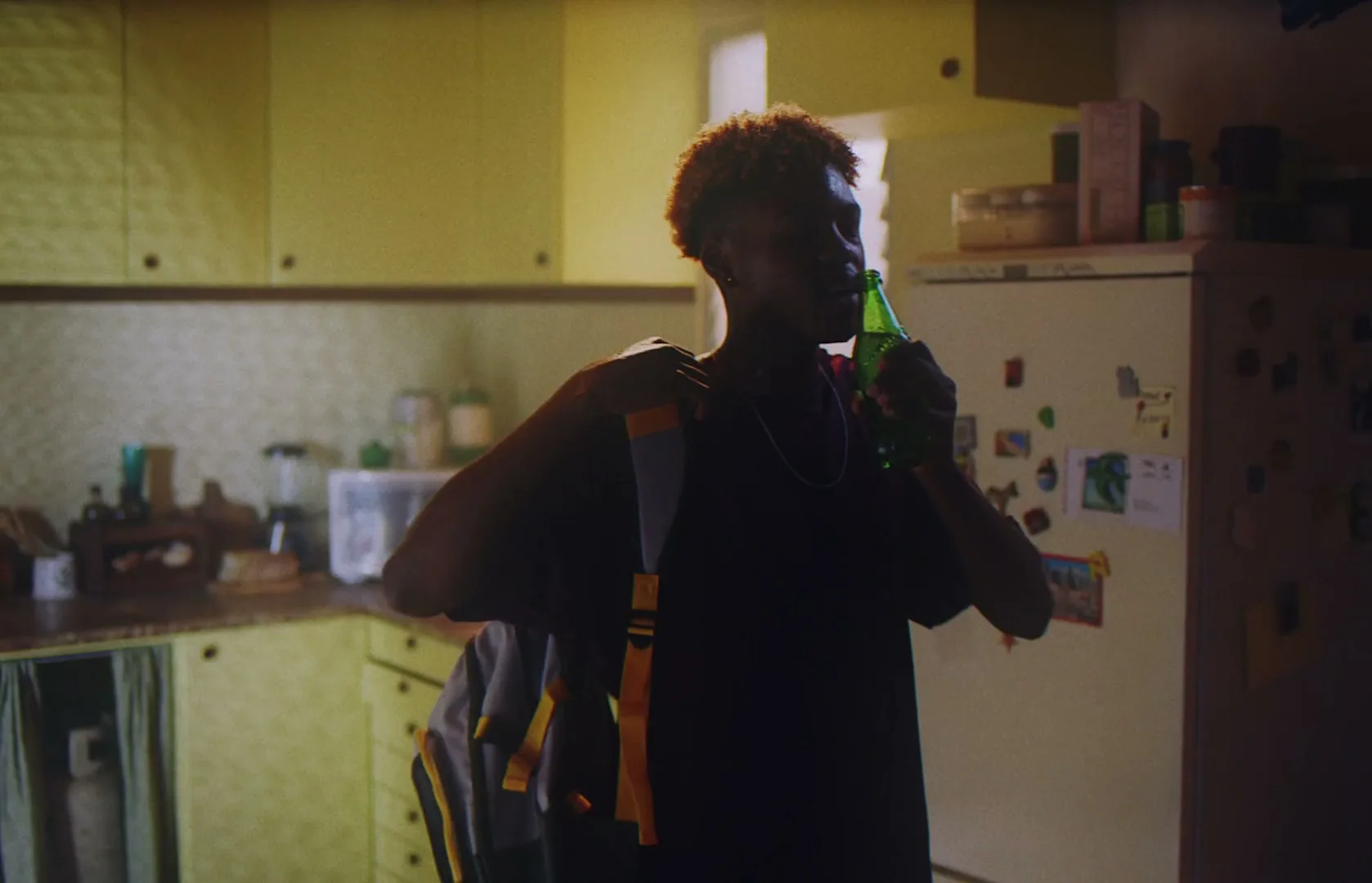 A young person with short, curly hair drinks from a green bottle in a dimly lit kitchen, then drops their backpack on the floor.