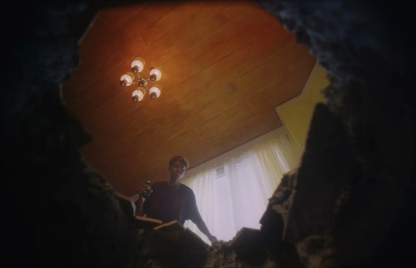 A young man in a dark t-shirt holds a green bottle, looking down into a hole in the floor of a room with a wooden ceiling and a chandelier.