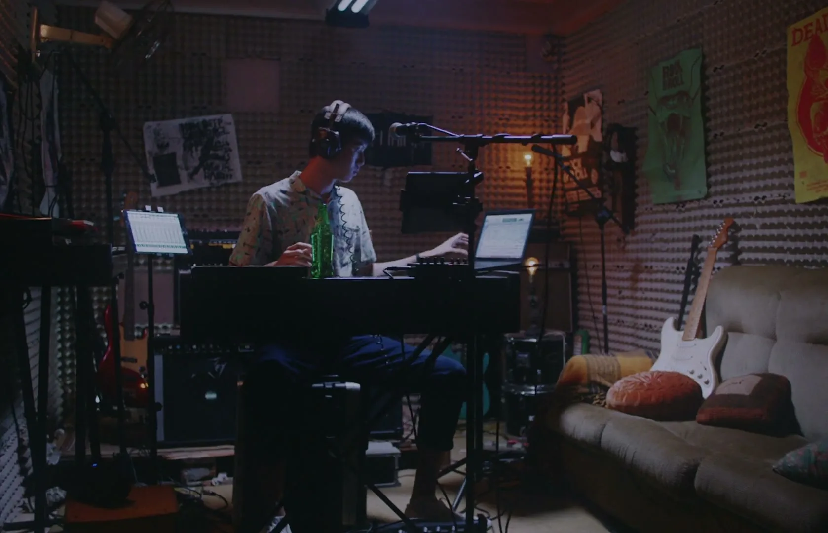 A young man in a patterned shirt and headphones sits at a keyboard in a dimly lit music studio, adjusting his headphones.
