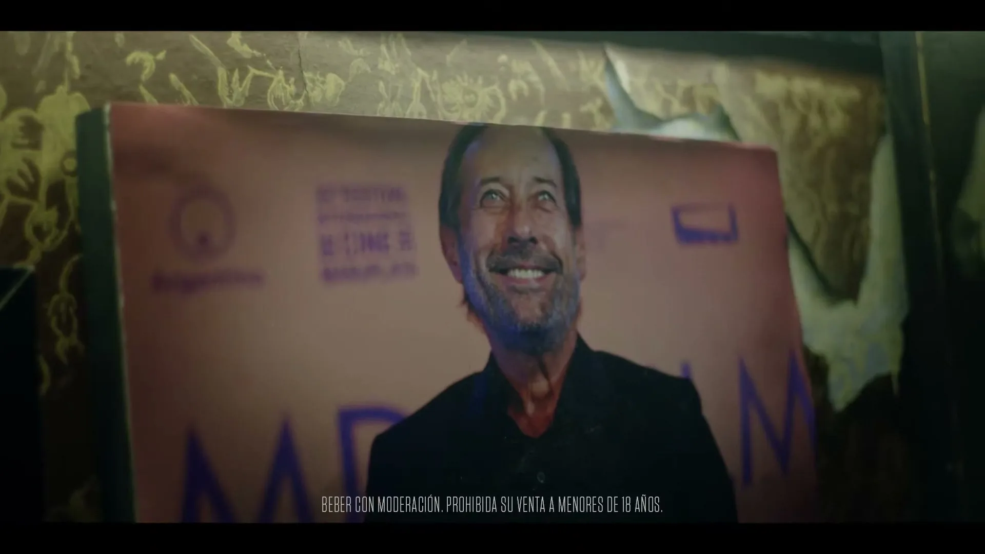 A poster of a smiling older man with a beard is shown on a wall with peeling wallpaper, with text at the bottom.