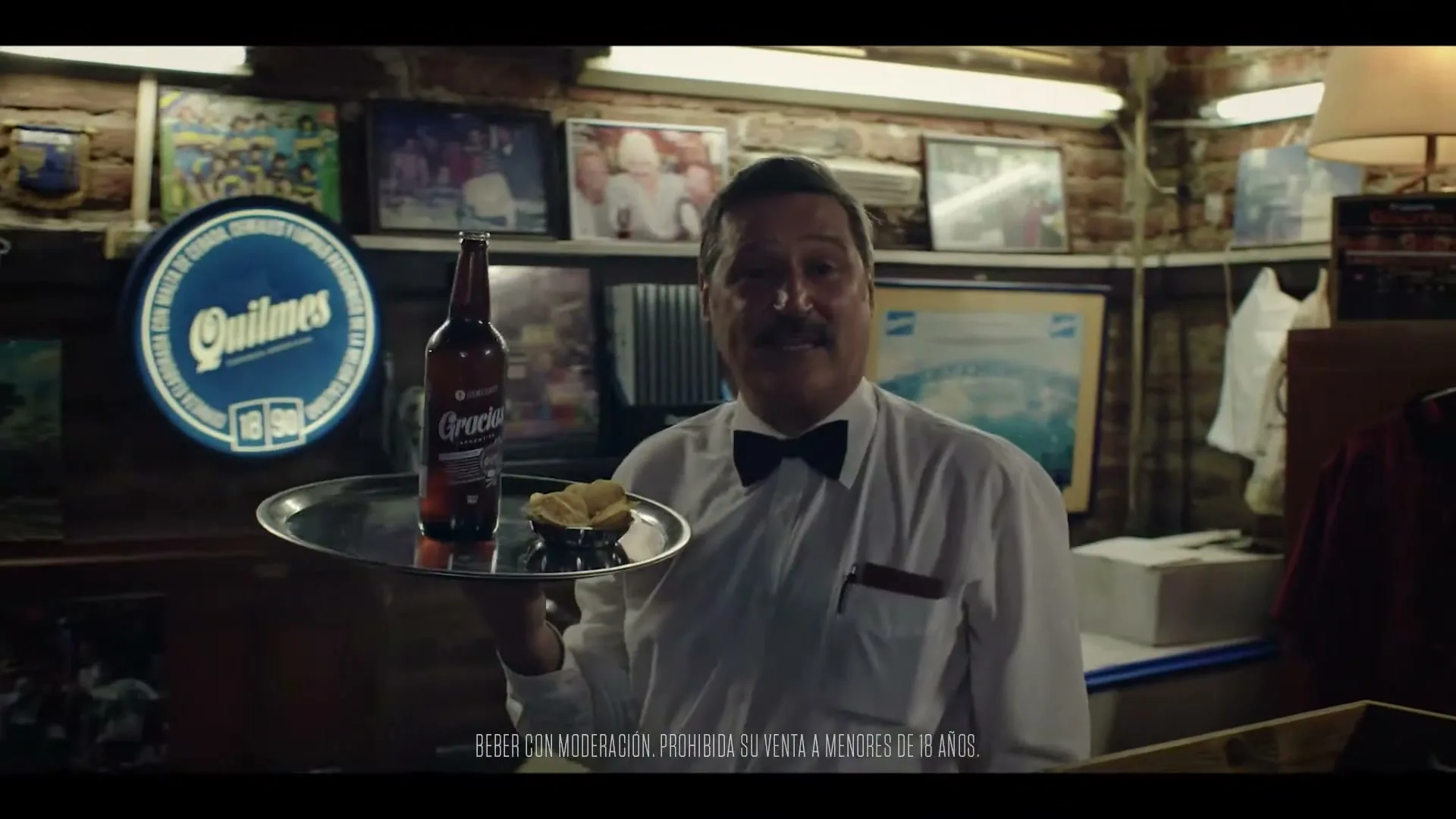 A smiling male waiter with a mustache holds a tray with a beer bottle and snacks in a dimly lit, rustic bar.
