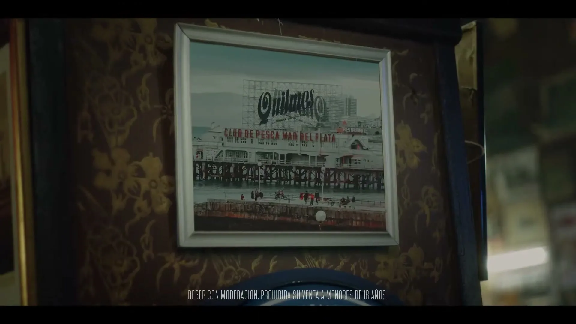 A framed picture of a pier with a large "Quilmes" sign hangs on a patterned wall, with legal text at the bottom of the screen.