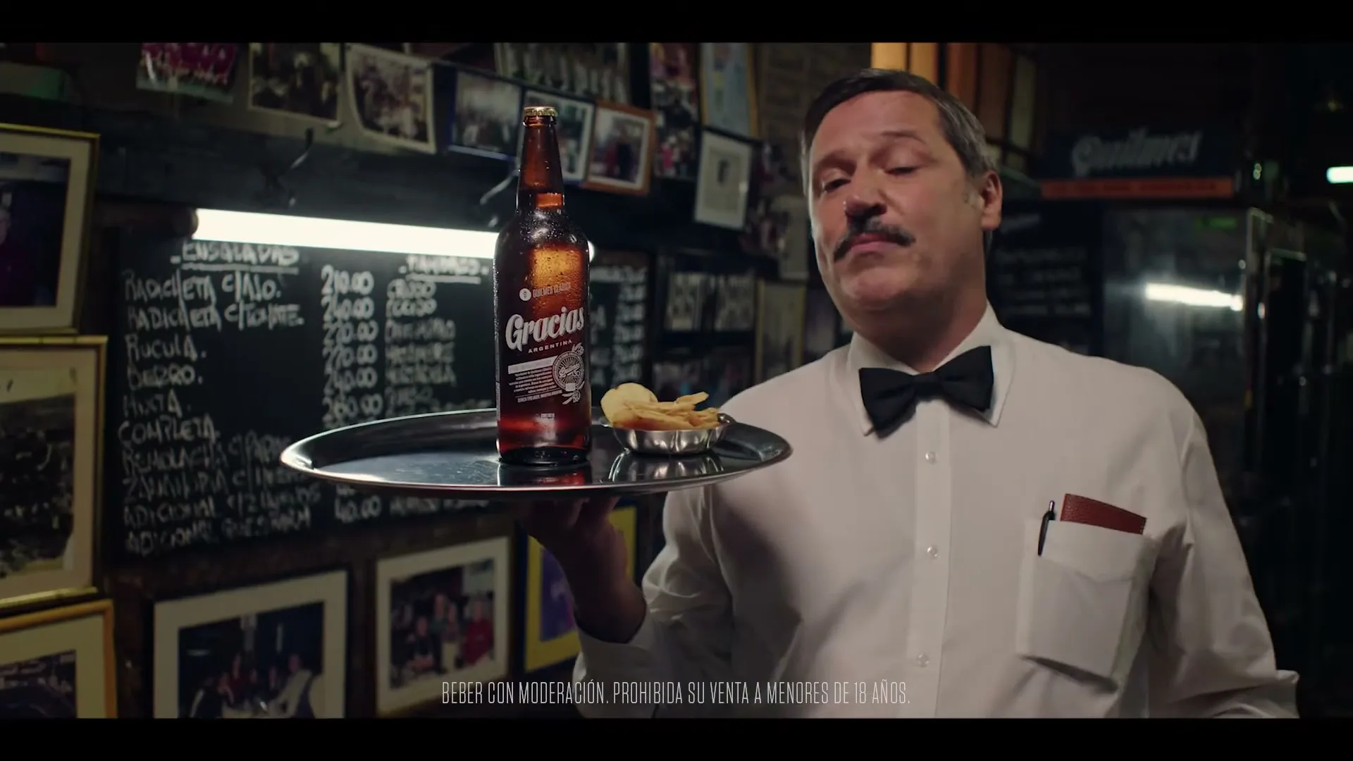 A male waiter with a mustache holds a tray with a beer bottle and chips, speaking directly to the camera in a dimly lit restaurant.
