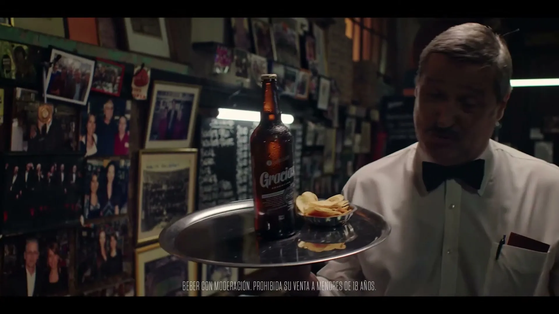 A male waiter with a mustache and bow tie holds a tray with a beer bottle and chips, looking down in a dimly lit bar.