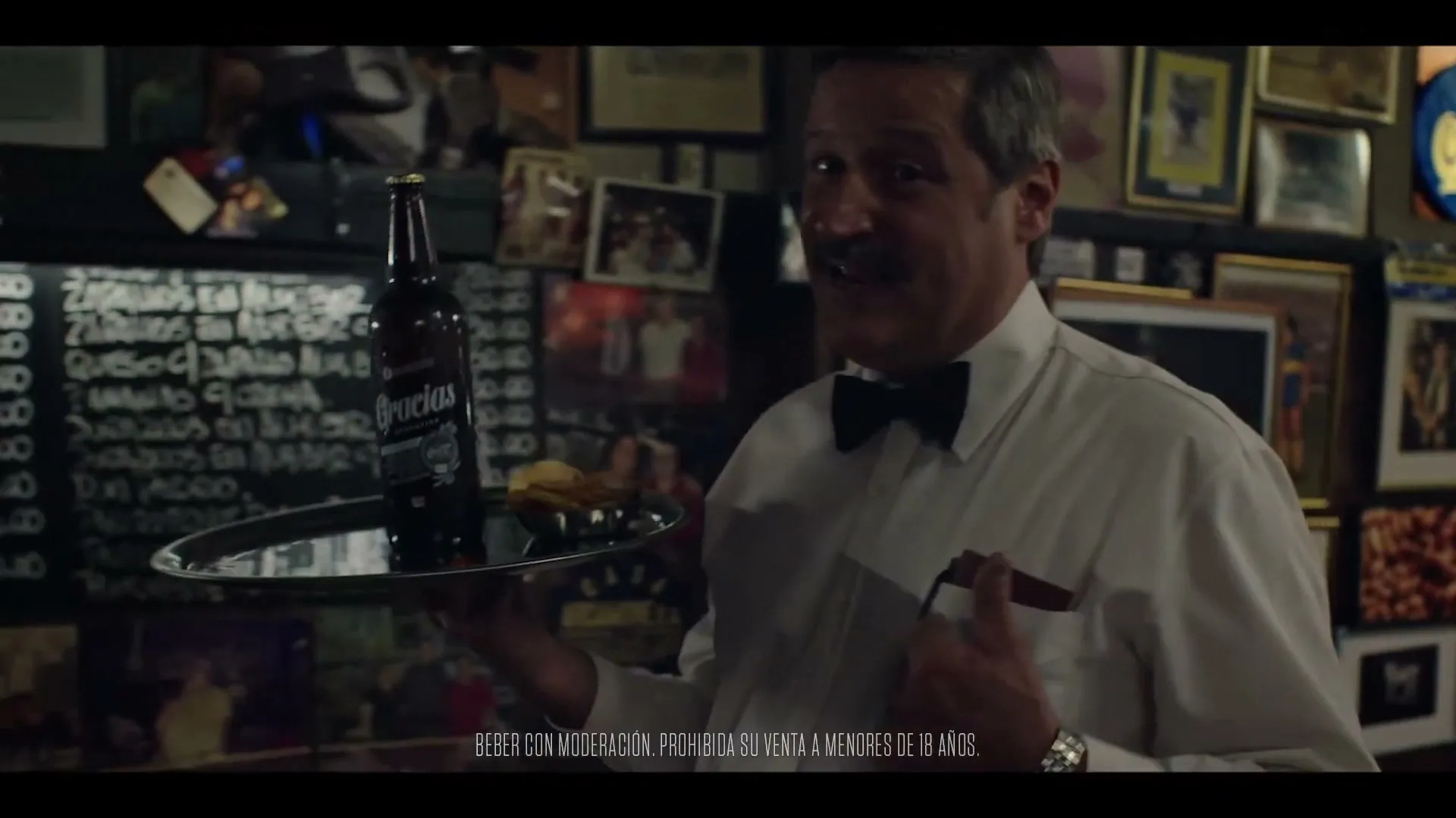 A male waiter in his 50s, wearing a white shirt and bow tie, holds a tray with a beer bottle and snacks in a dimly lit bar.