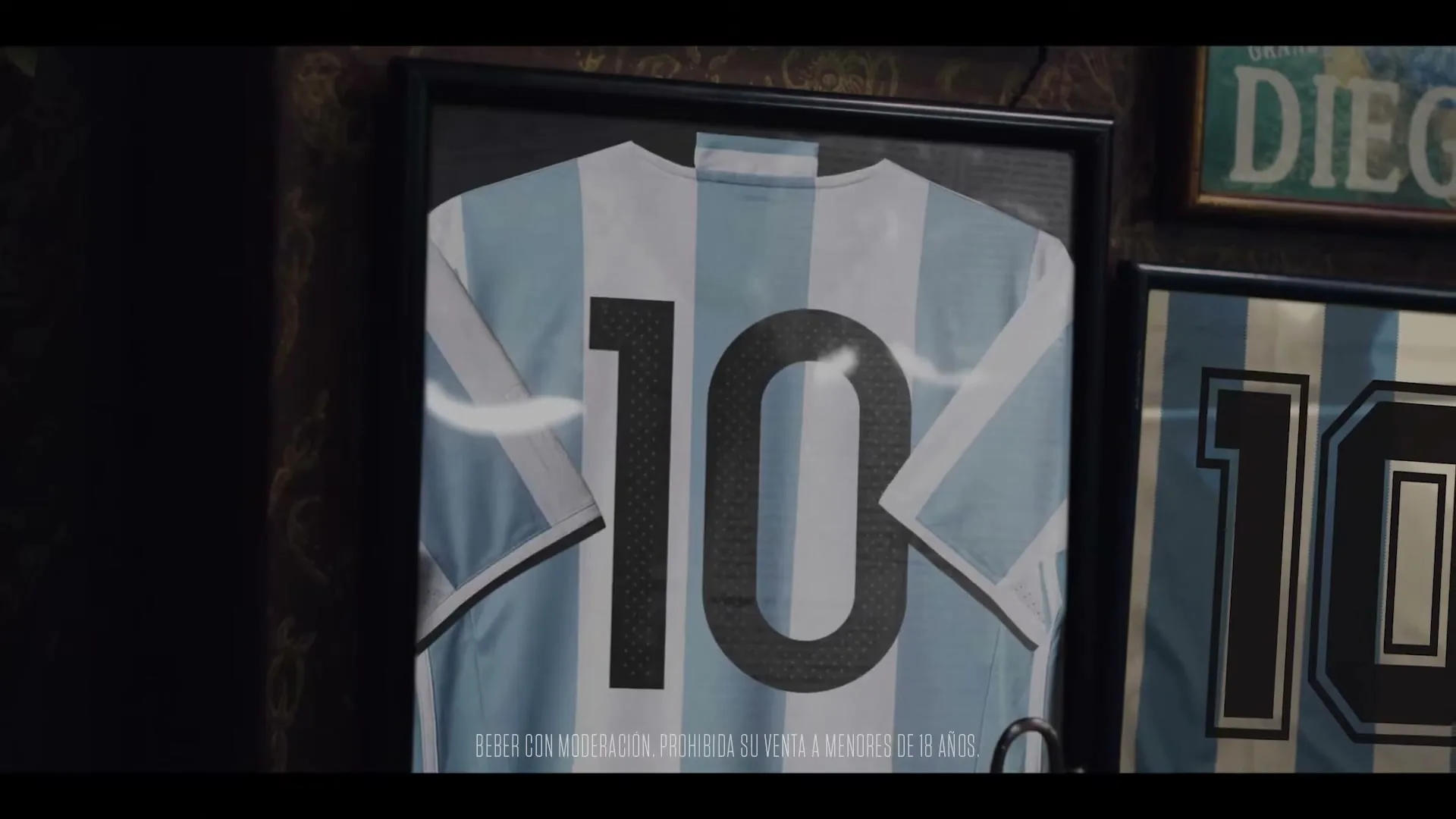 A framed light blue and white striped soccer jersey with the number 10 on the back hangs on a dark wall, with a glimpse of another jersey and a "DIEGO" sign.