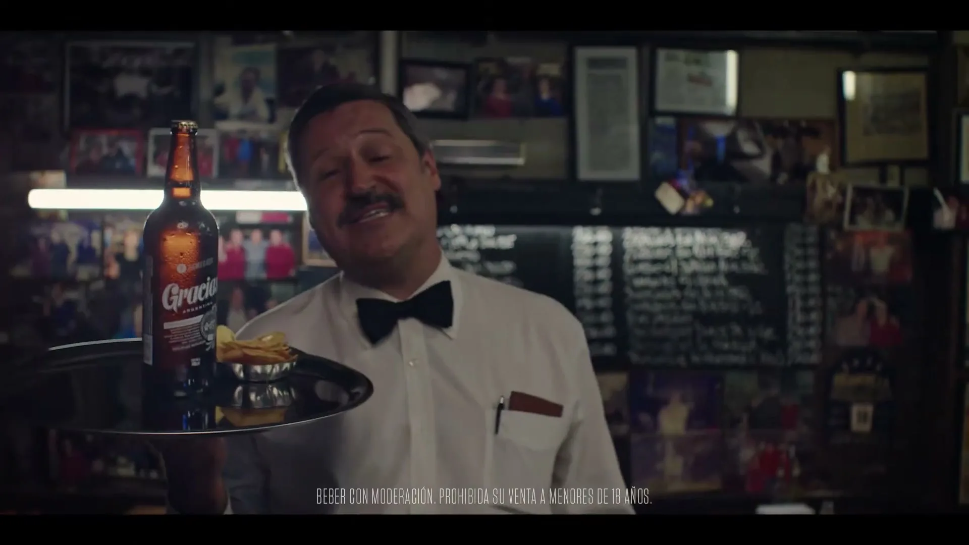A male waiter with a mustache, wearing a white shirt and bow tie, holds a tray with a beer bottle and snacks in a dimly lit bar.