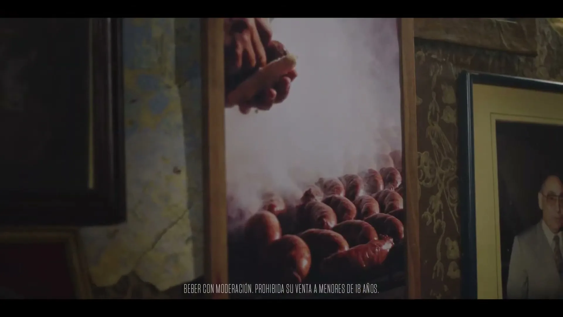 A framed photograph of sausages cooking on a grill with smoke rising is displayed on a wall, next to another framed portrait.