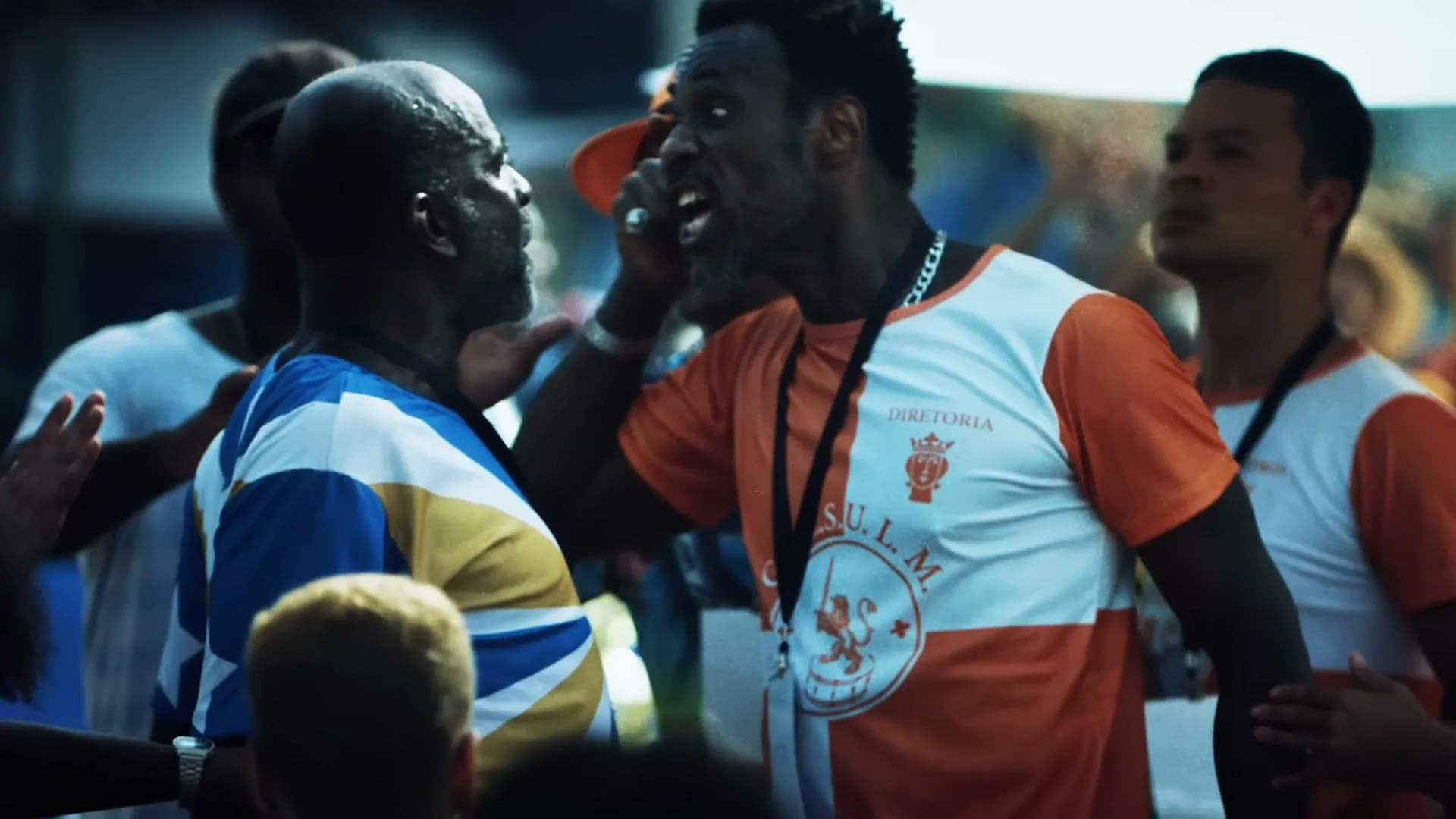 Two men, one in a blue and yellow shirt and another in an orange and white shirt, are arguing intensely in a crowded outdoor setting.