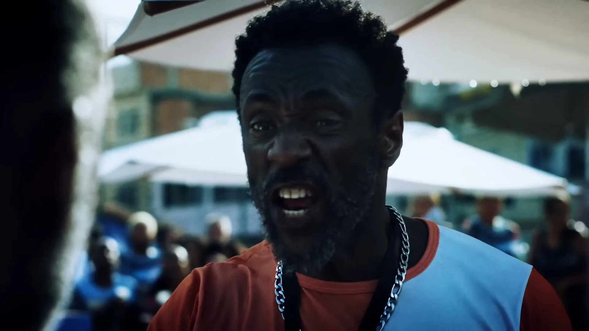 A dark-skinned man with a beard and short curly hair, wearing an orange and white shirt and a silver chain, shouts at someone off-camera in an outdoor setting.
