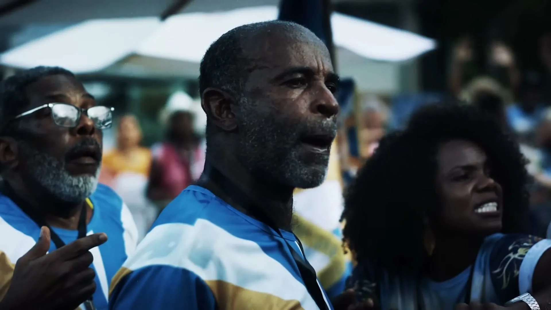 A diverse group of people, including a man with a grey beard and a woman with curly hair, are singing and dancing at an outdoor carnival.